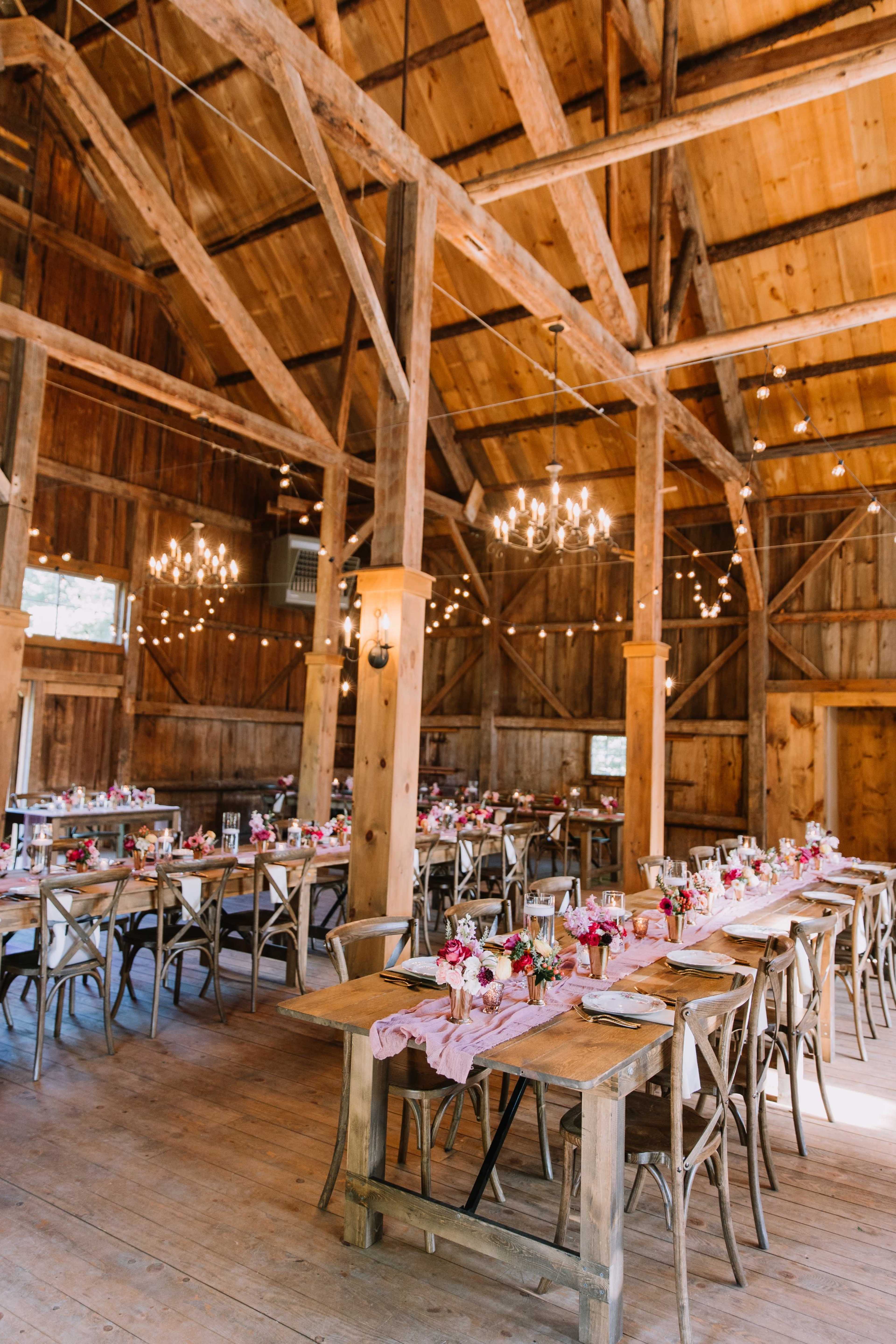 The interior of a rustic barn features long dining tables with floral arrangements, wooden beams, and chandeliers.