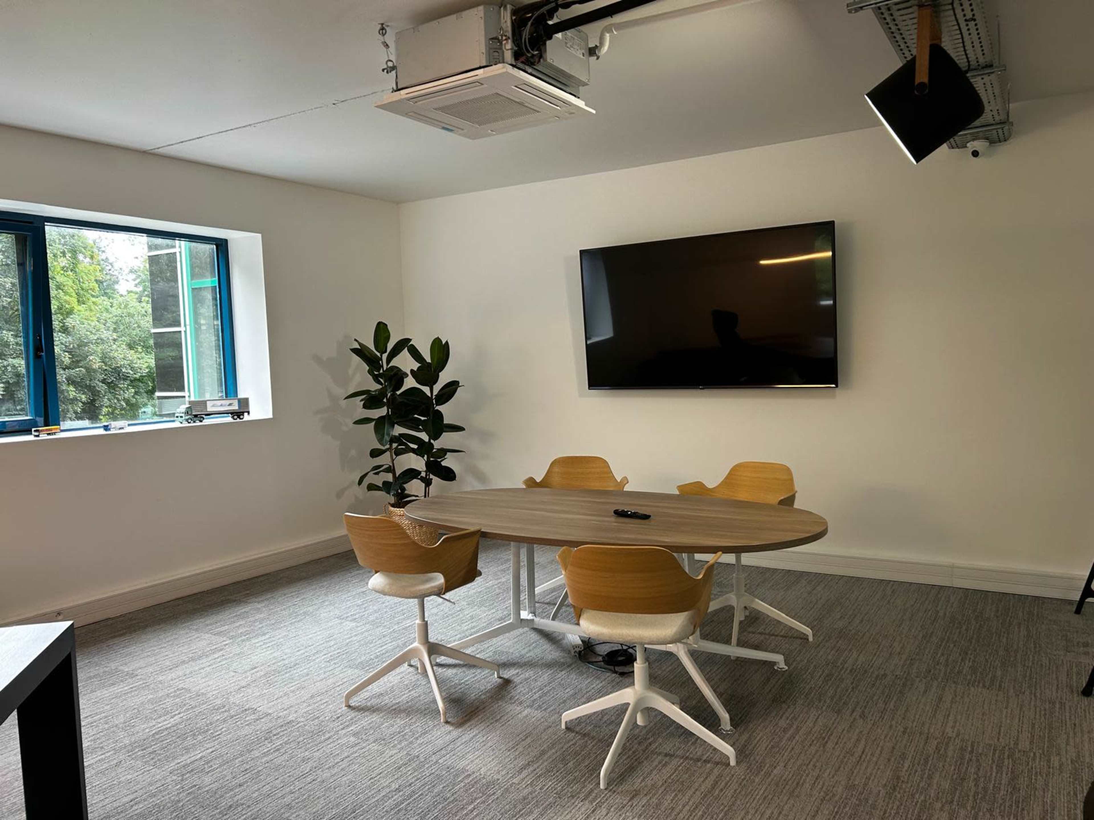 The image shows a modern meeting room with a round wooden table, four chairs, a large wall-mounted screen, and a potted plant near a window.