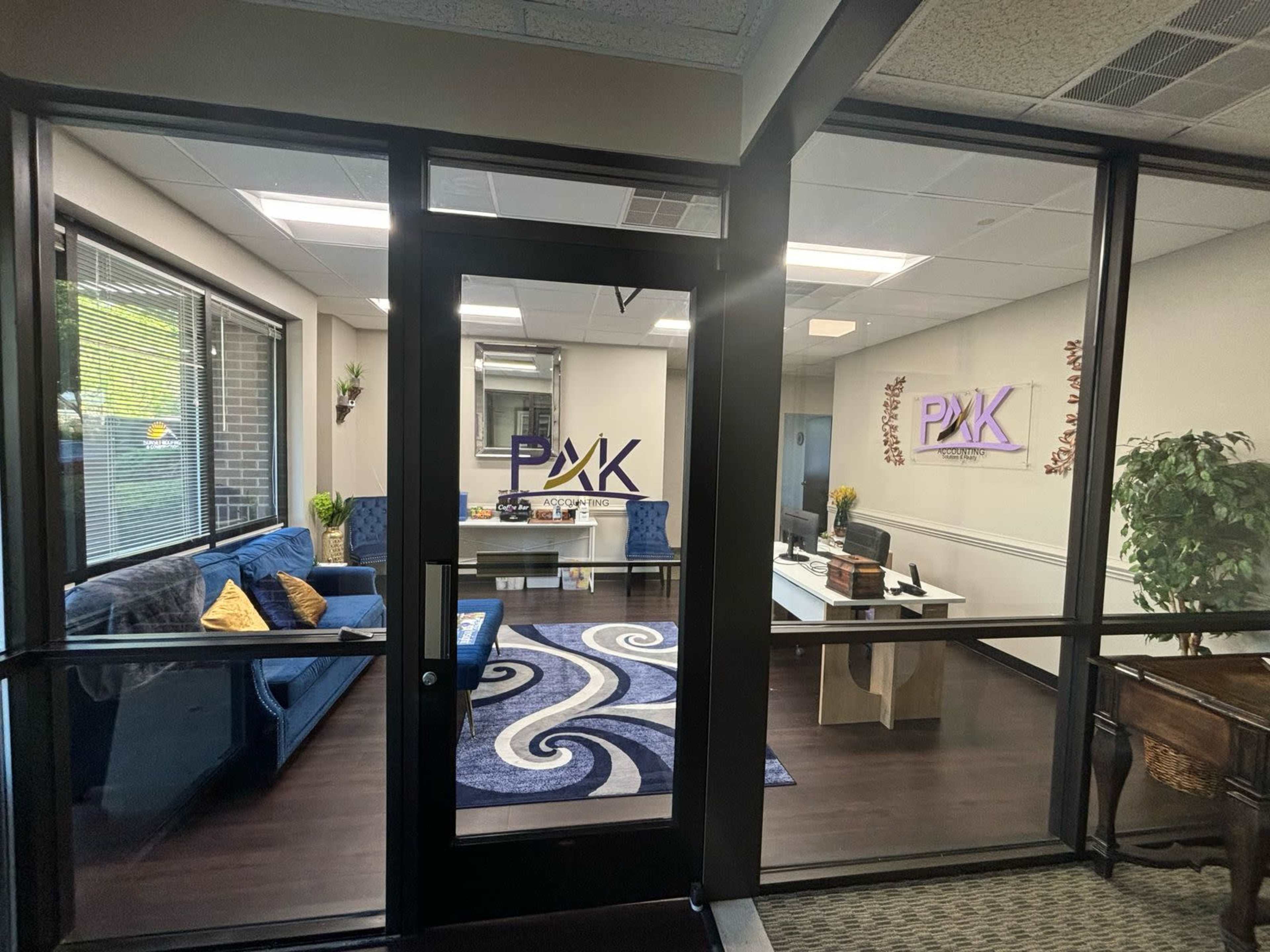 The image shows a modern office space with a reception area featuring blue couches, a patterned rug, and a desk with a television visible through glass doors.