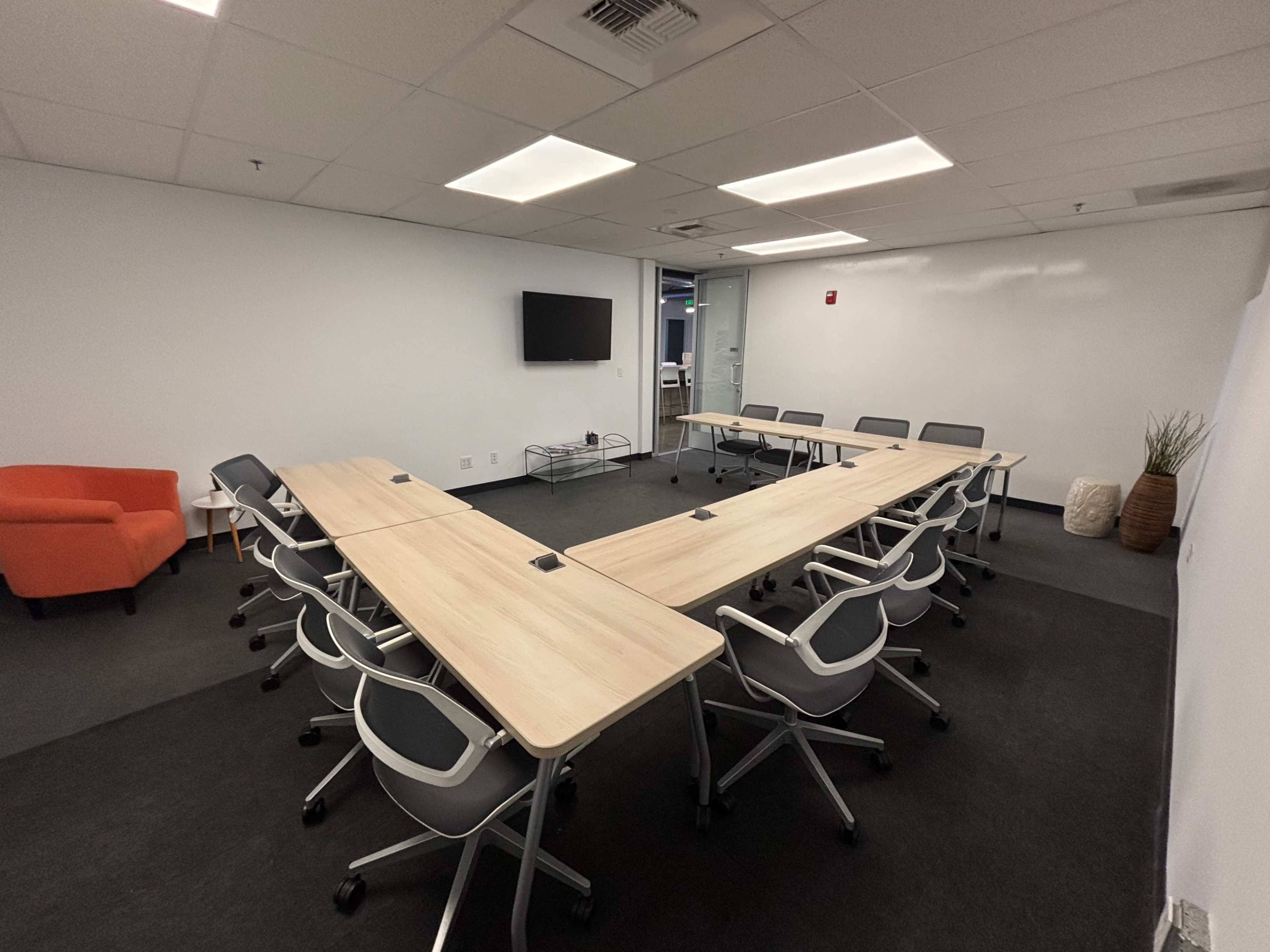 The image shows a conference room with a long table surrounded by chairs, a television mounted on the wall, and an orange armchair in one corner.