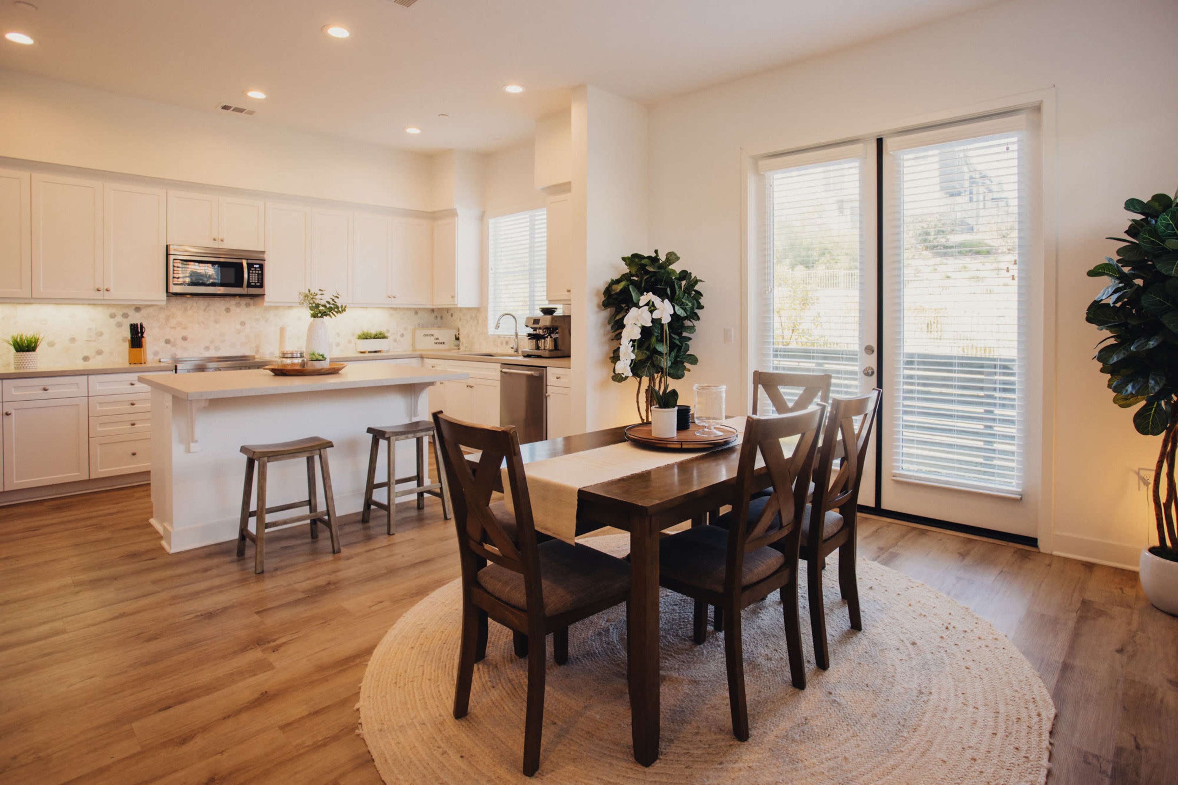 A modern kitchen and dining area features a wooden table with six chairs, a light-colored rug, and a window facing a garden.