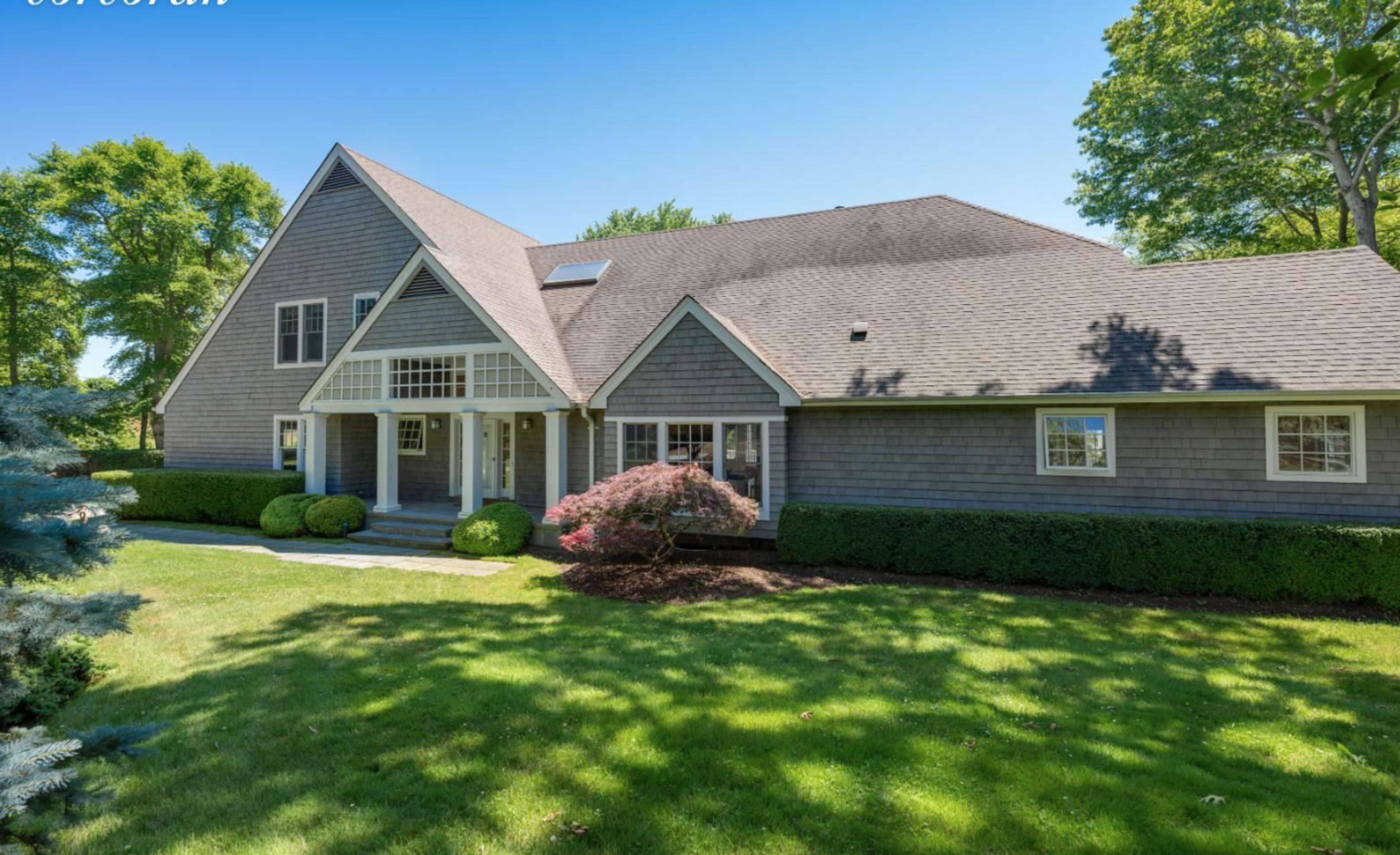 A large gray shingle-style house with a gabled roof is surrounded by a well-maintained lawn and mature trees.