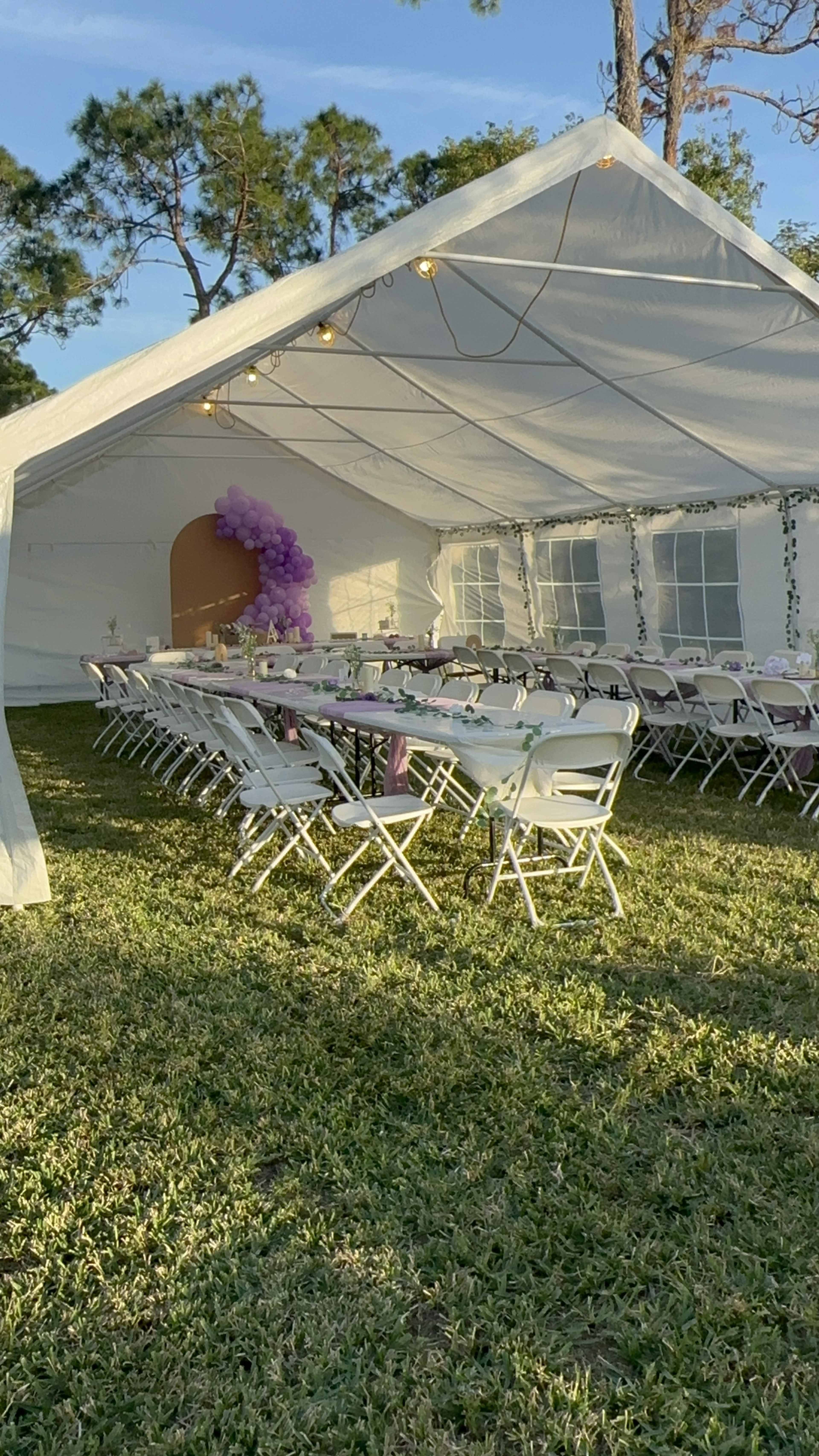 A white tent with tables set for an event is arranged on a grassy area, featuring decorative touches of purple and string lights.
