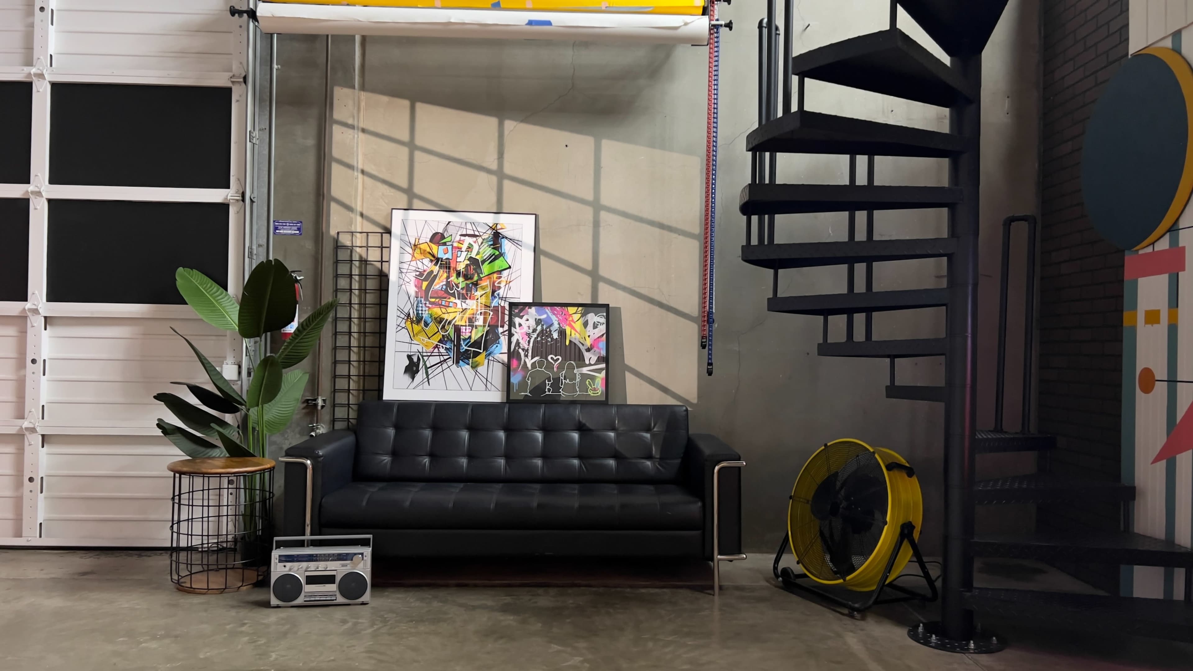 The image features a black leather sofa positioned next to two vibrant artworks on a wall, a spiral staircase in the background, and a yellow fan on the floor beside a potted plant.