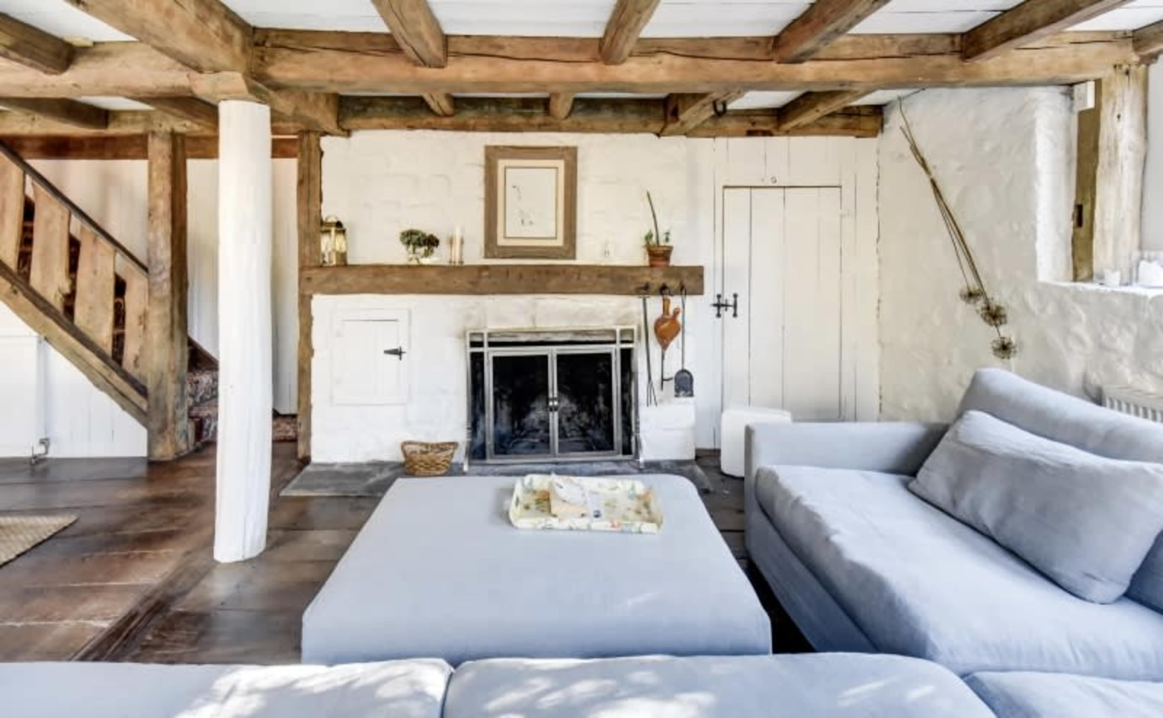 A cozy living room with exposed wooden beams, a stone fireplace, and a light-colored sofa arranged around a square coffee table.