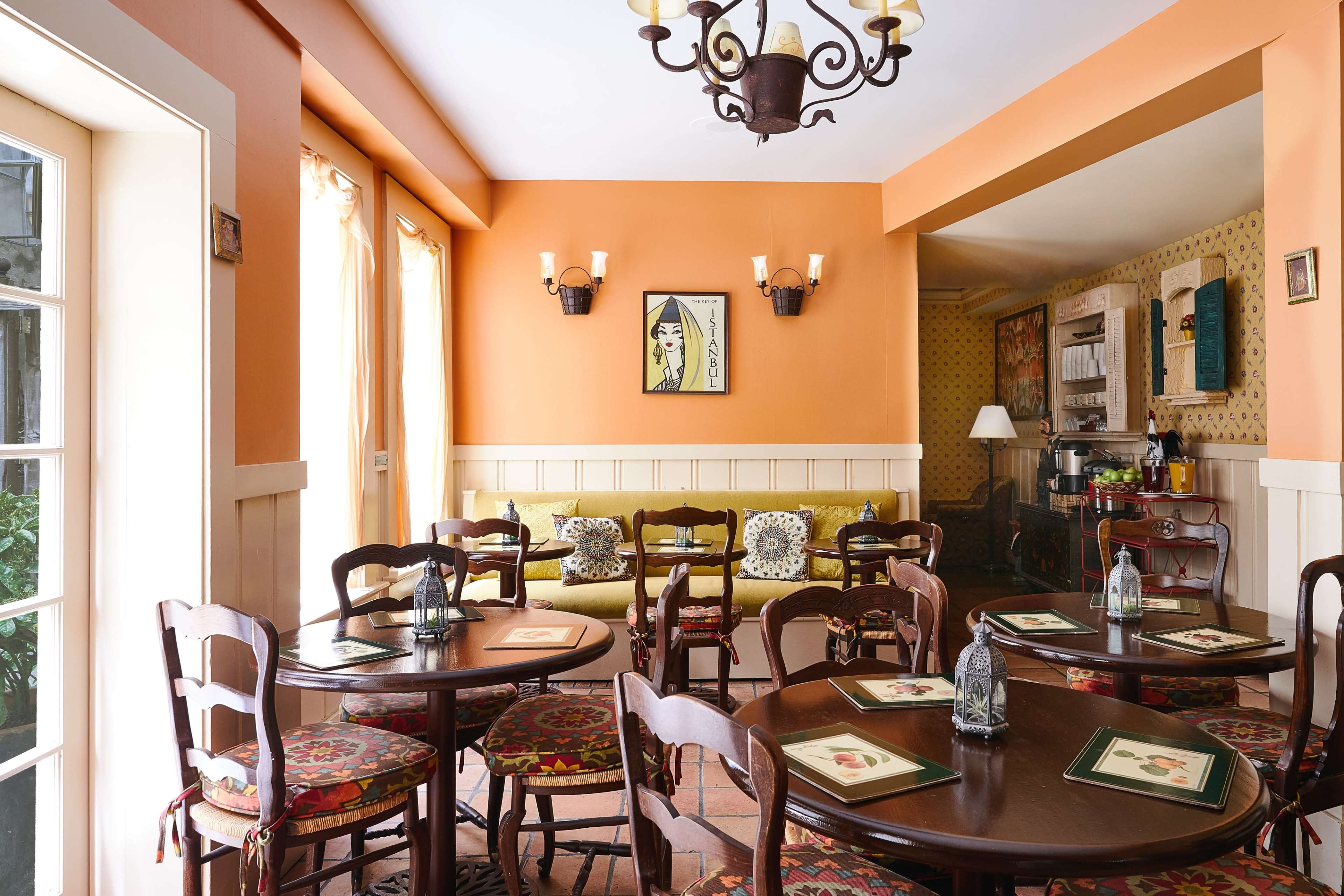A cozy dining area with brown wooden tables and chairs, an orange wall, and a small seating nook against the wall, illuminated by natural light.