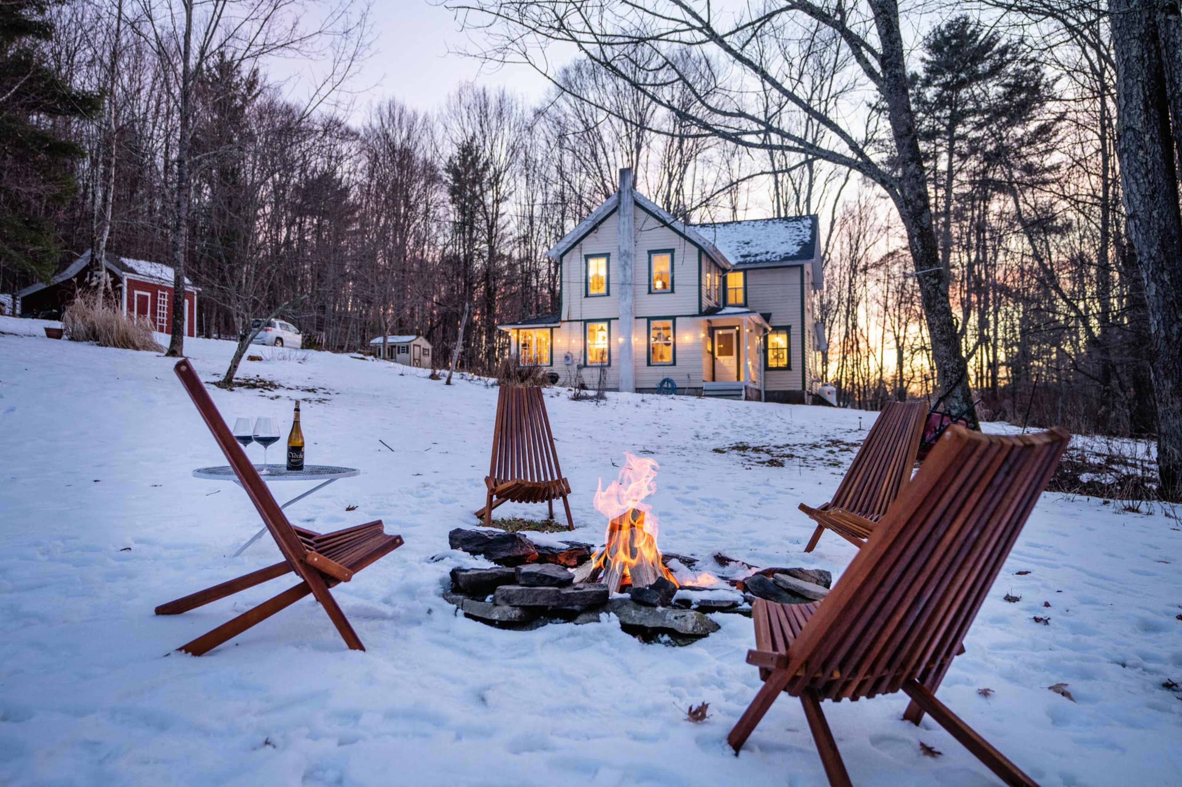 A fire pit surrounded by six wooden chairs sits in the snow, with a house in the background and a sunset illuminating the scene.