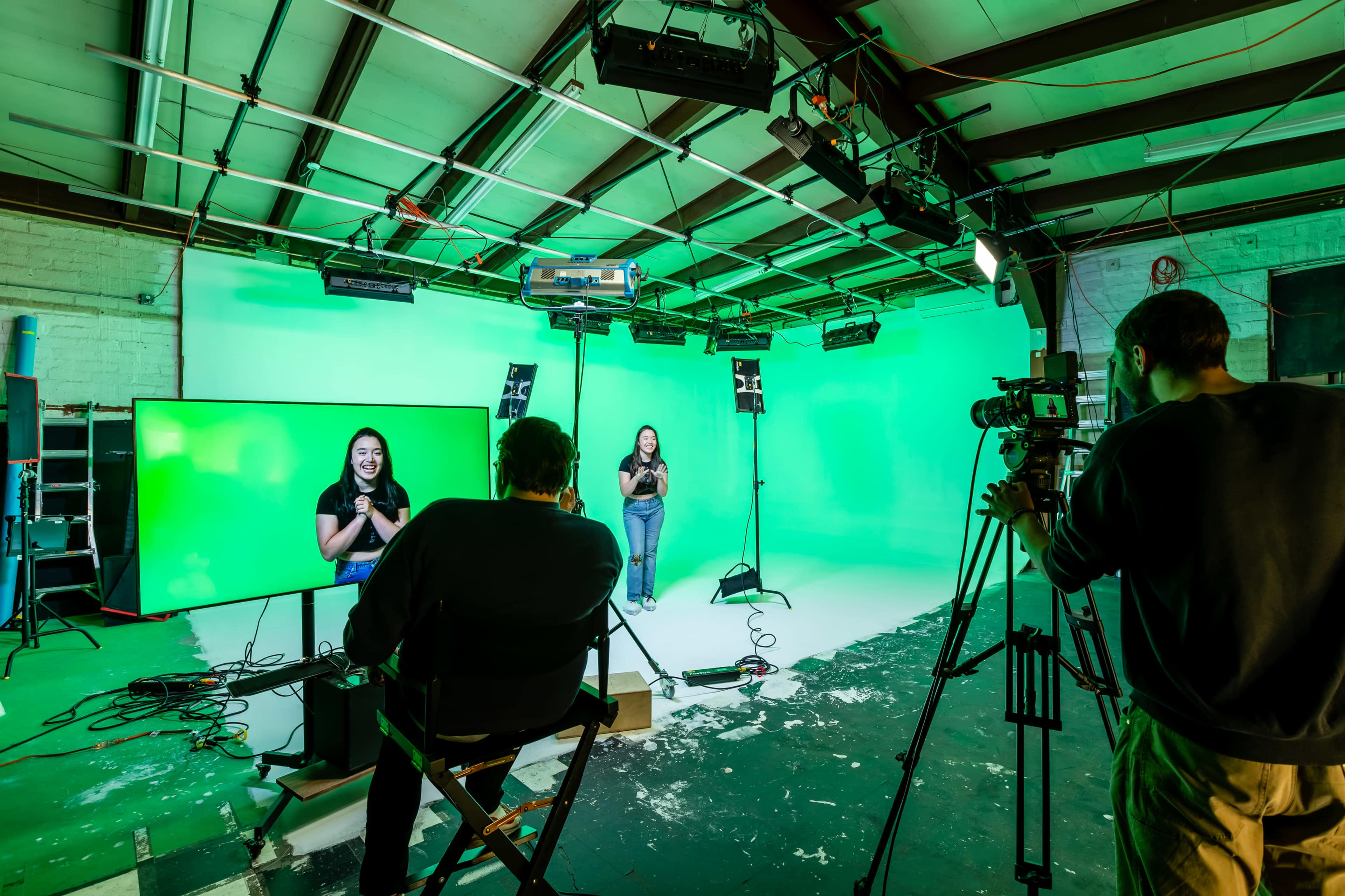 A person stands in front of a green screen while another individual films from a tripod in a studio setting.