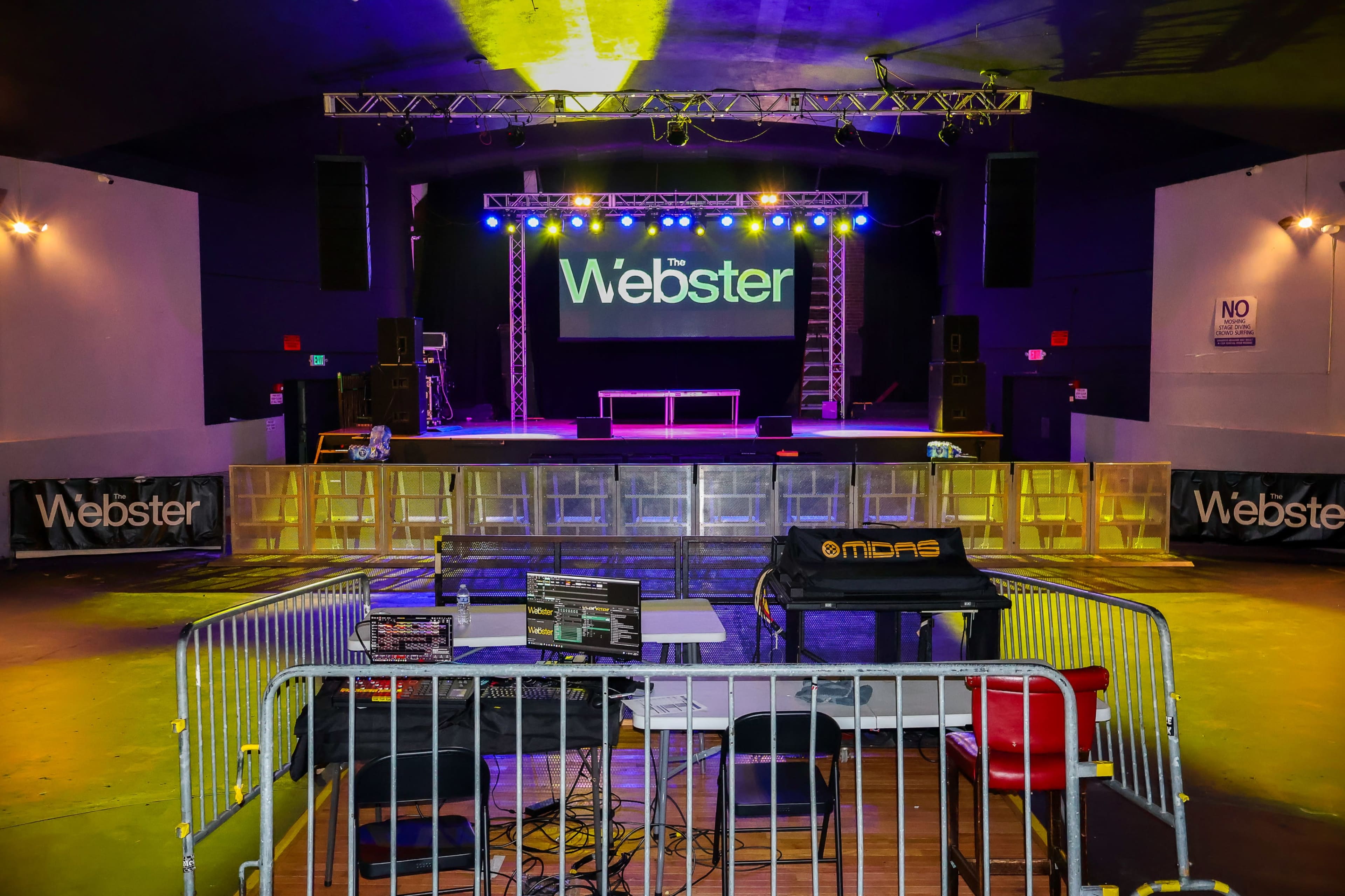 The image shows a music venue stage with lighting set up, featuring a large "Webster" sign and barriers separating the audience area from the stage equipment.