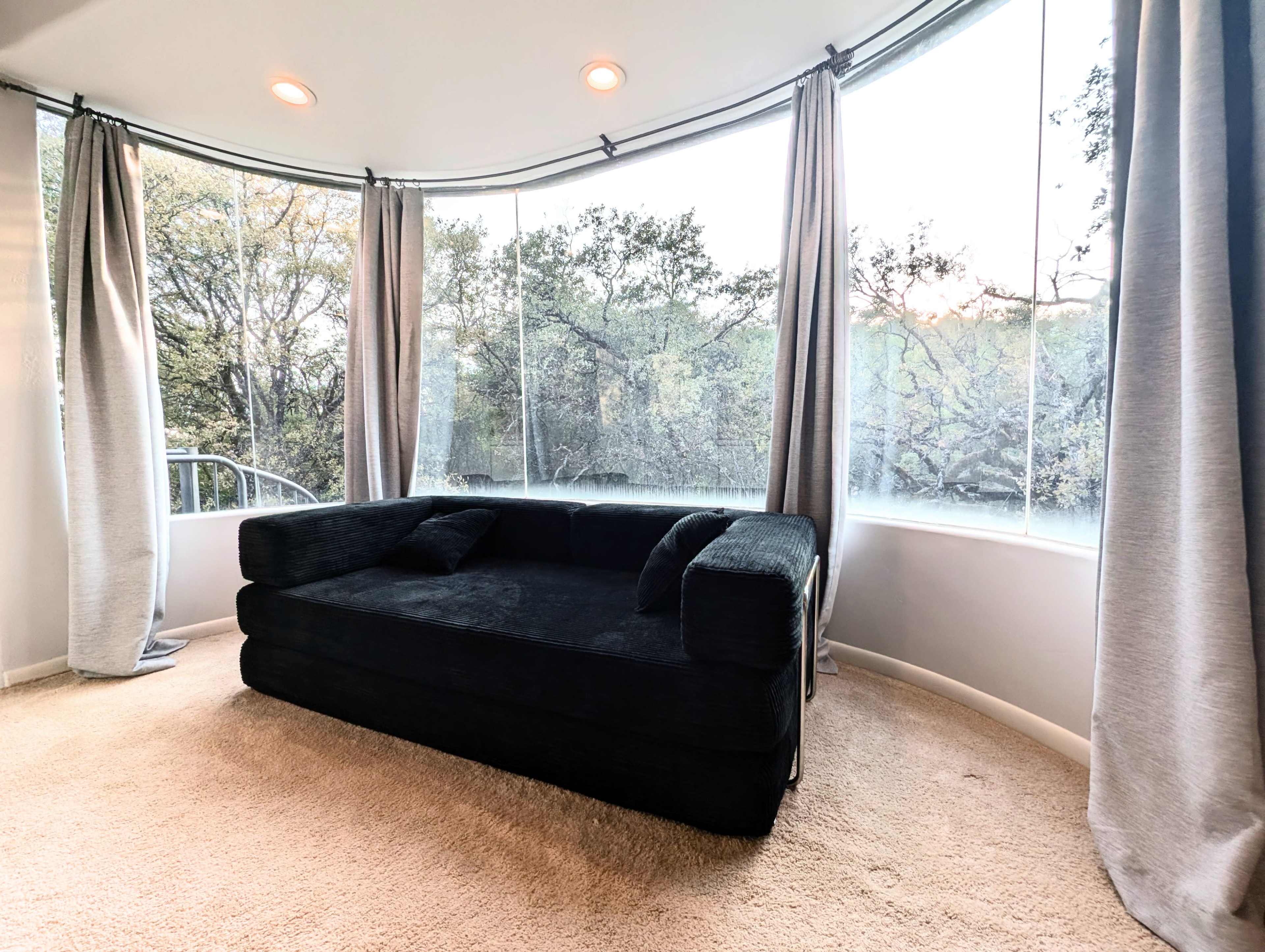The image shows a black sectional sofa positioned in a curved window alcove, surrounded by light curtains.