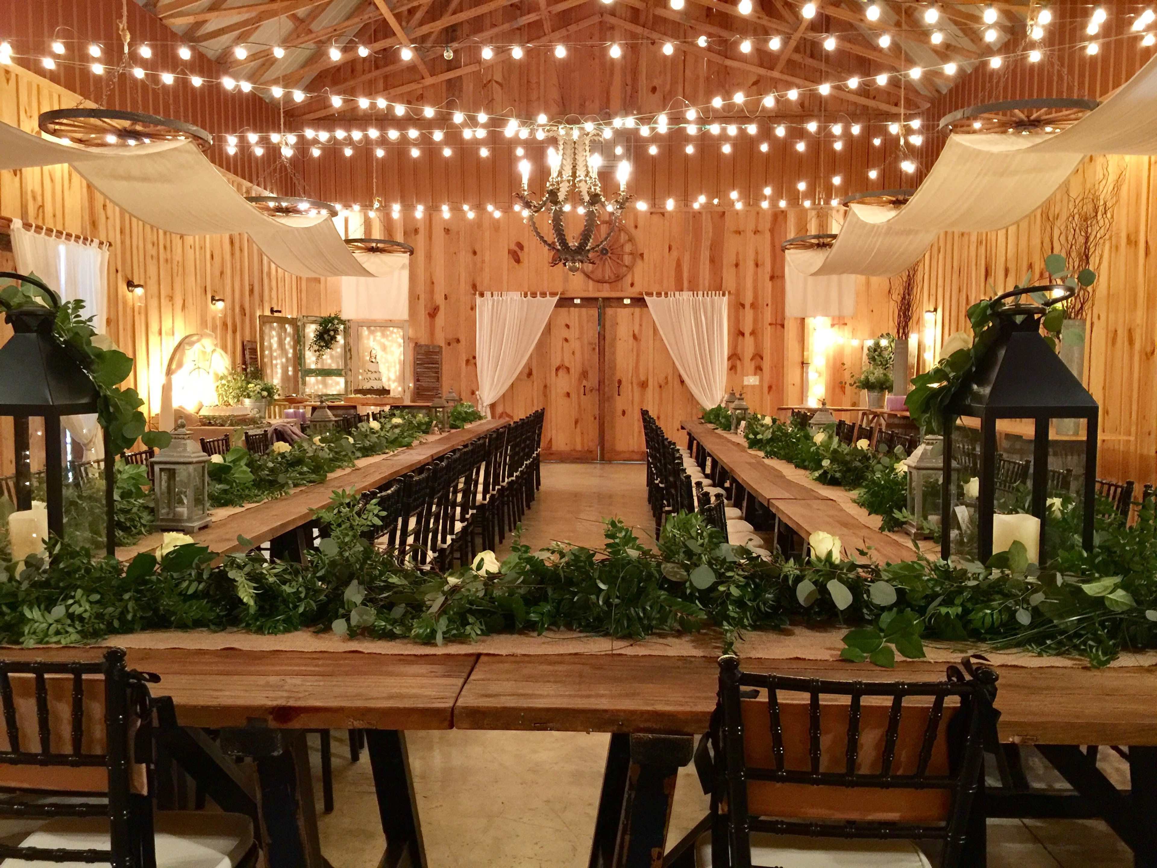 A spacious, wooden banquet hall is decorated with string lights and greenery along long wooden tables, with rows of dark chairs facing the center.
