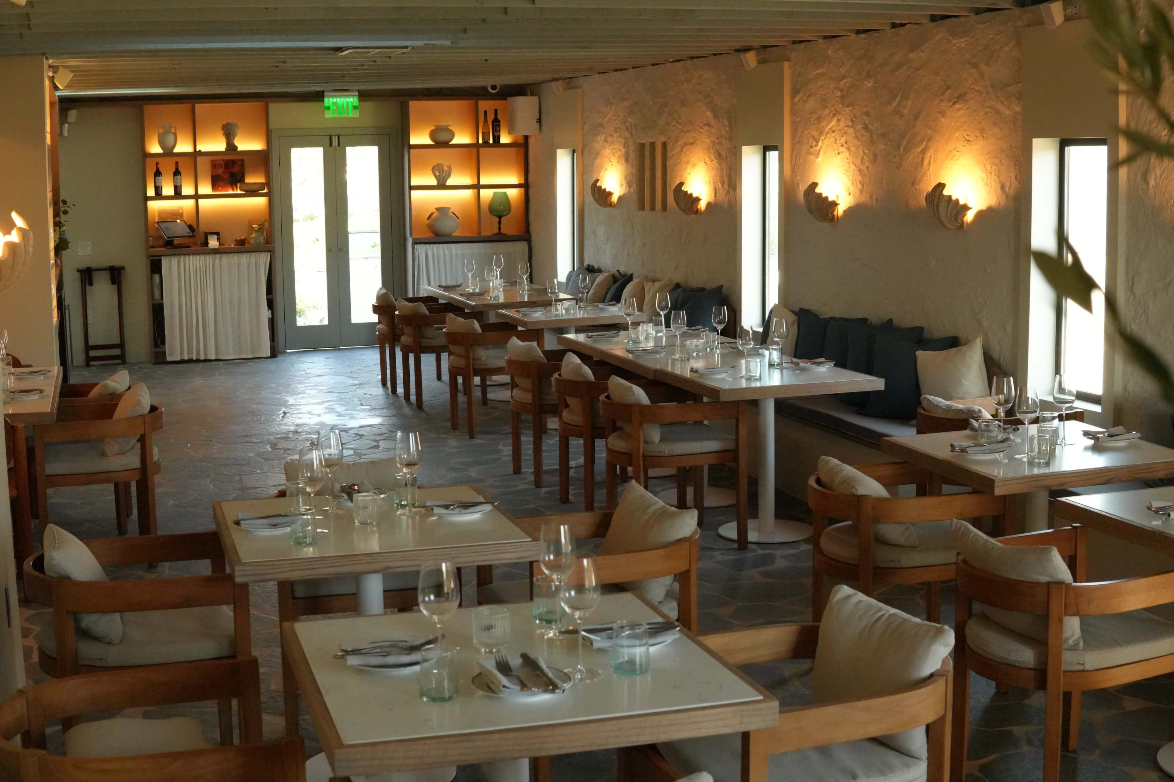 The image shows a modern restaurant interior featuring wooden tables, light-colored walls, and decorative shelves with various items.