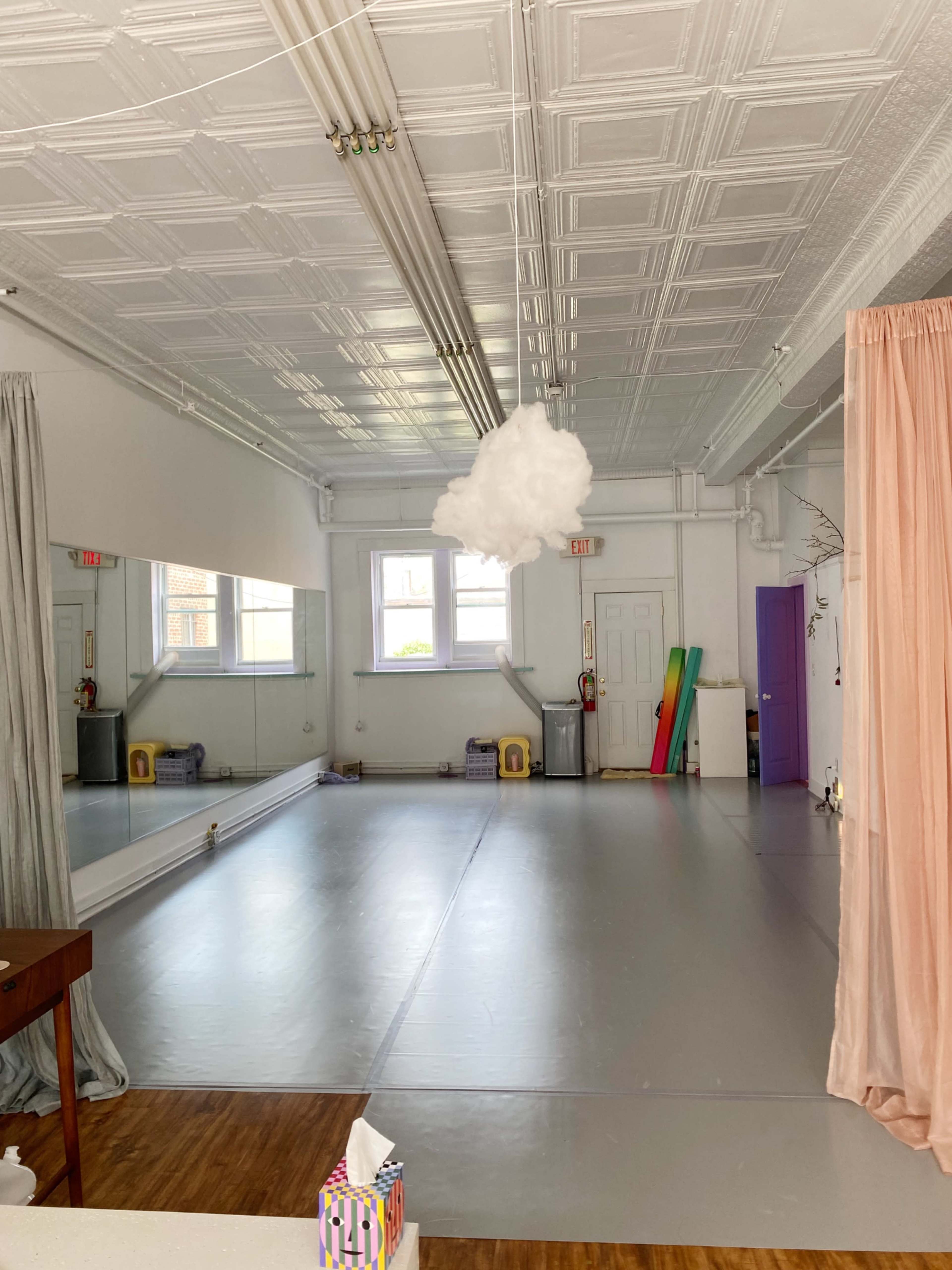 The image shows a spacious dance studio with a mirrored wall, light gray flooring, and a cloud-like decoration hanging from the ceiling.