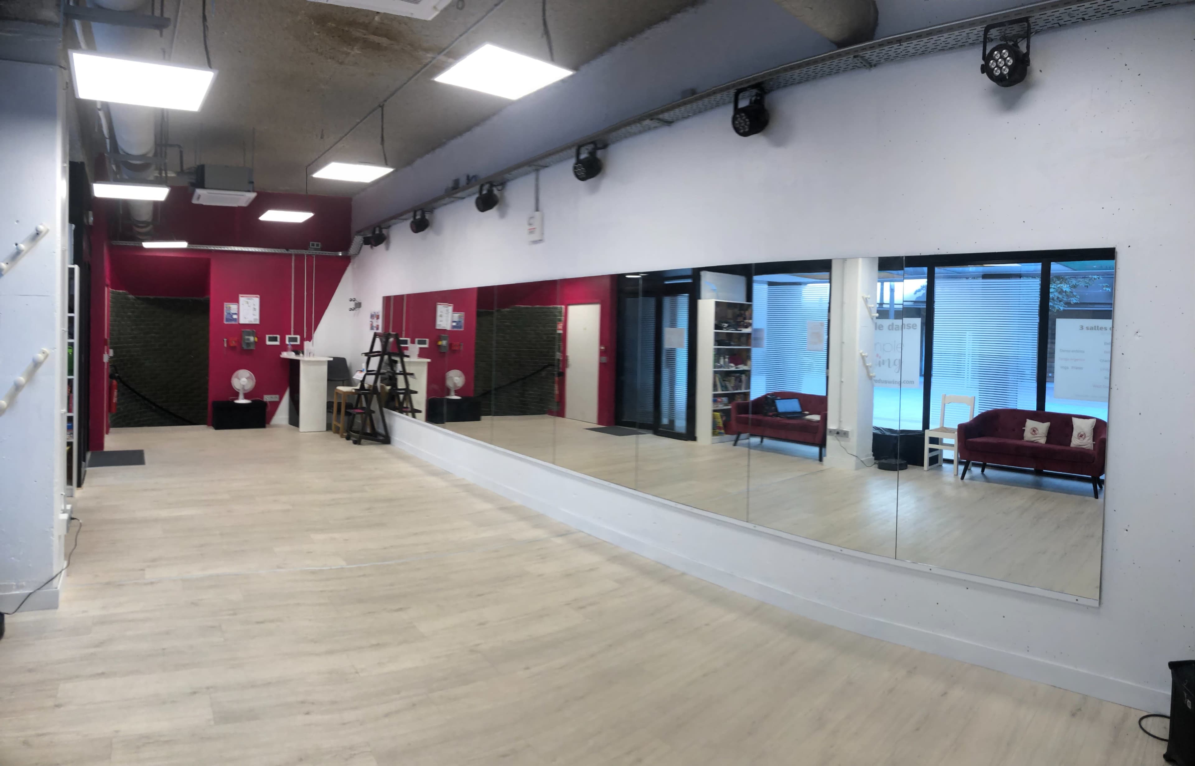 A spacious dance studio with wooden flooring, large mirrors on one wall, and pink and white walls, featuring a seating area with red sofas.