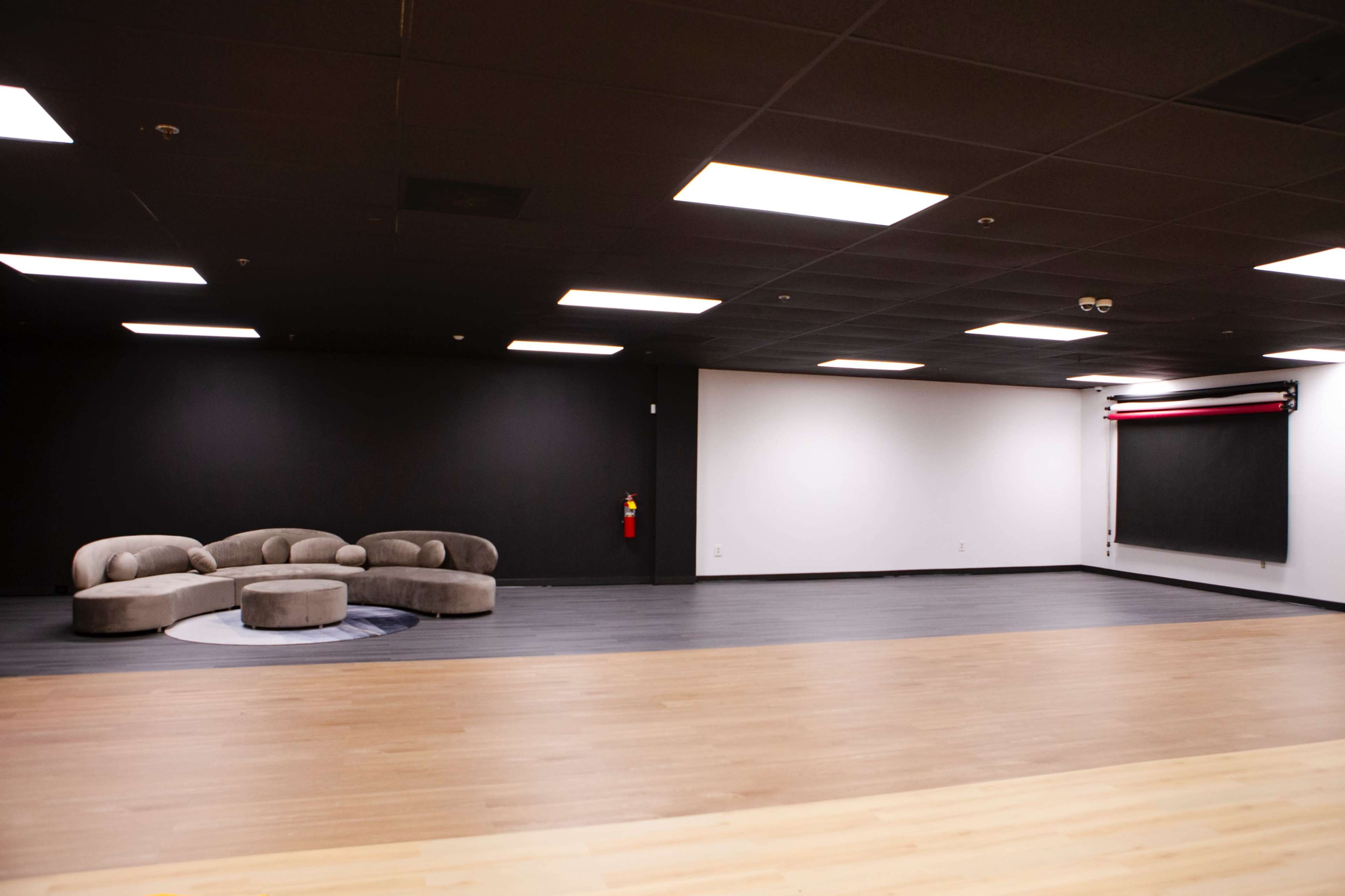 The image shows a spacious room with a circular gray sofa, a wooden floor, and a large black panel against one wall.