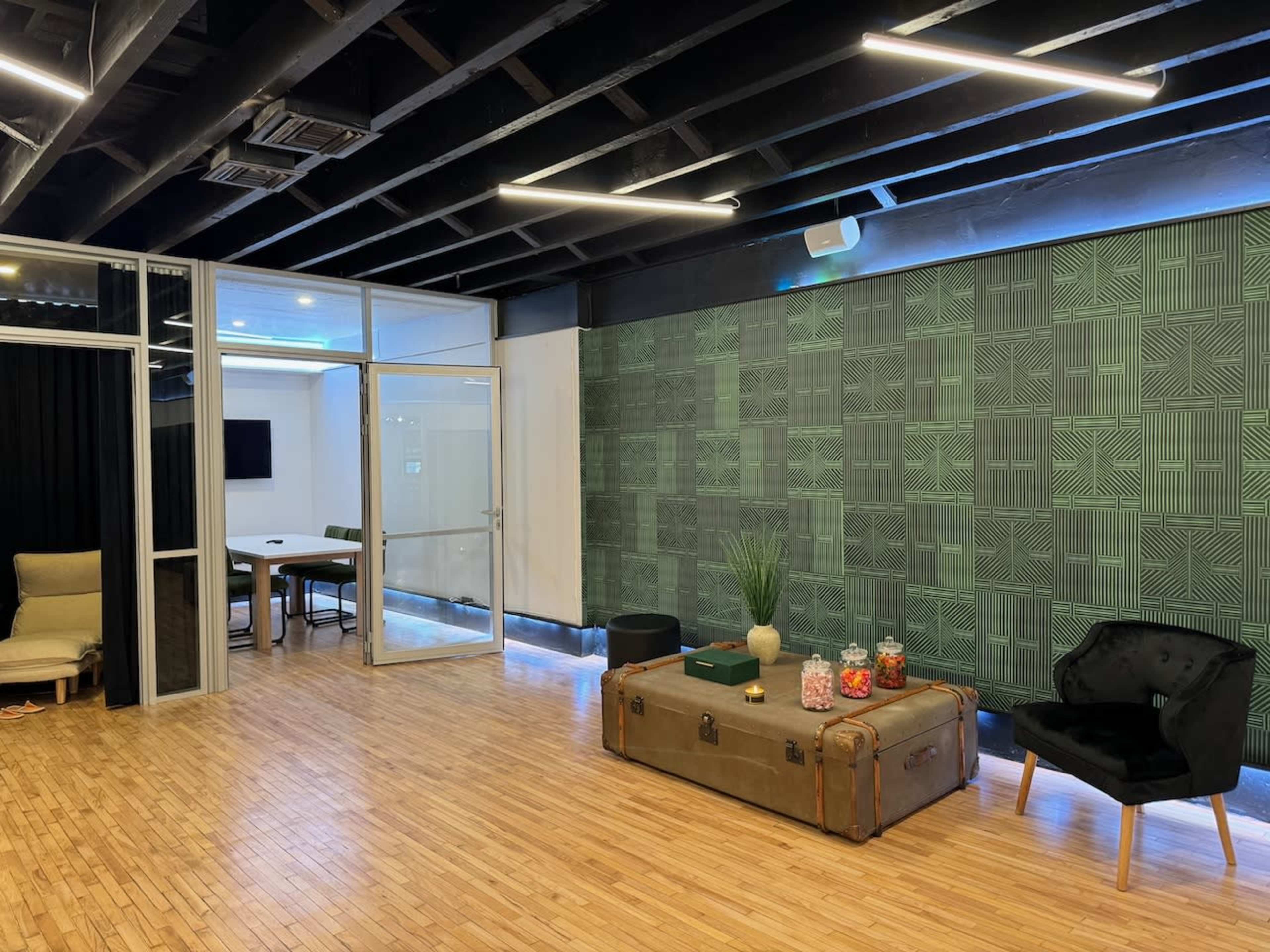 The image shows a modern office interior with a wooden floor, a seating area featuring a black chair and a vintage trunk, and a glass-walled meeting room in the background.