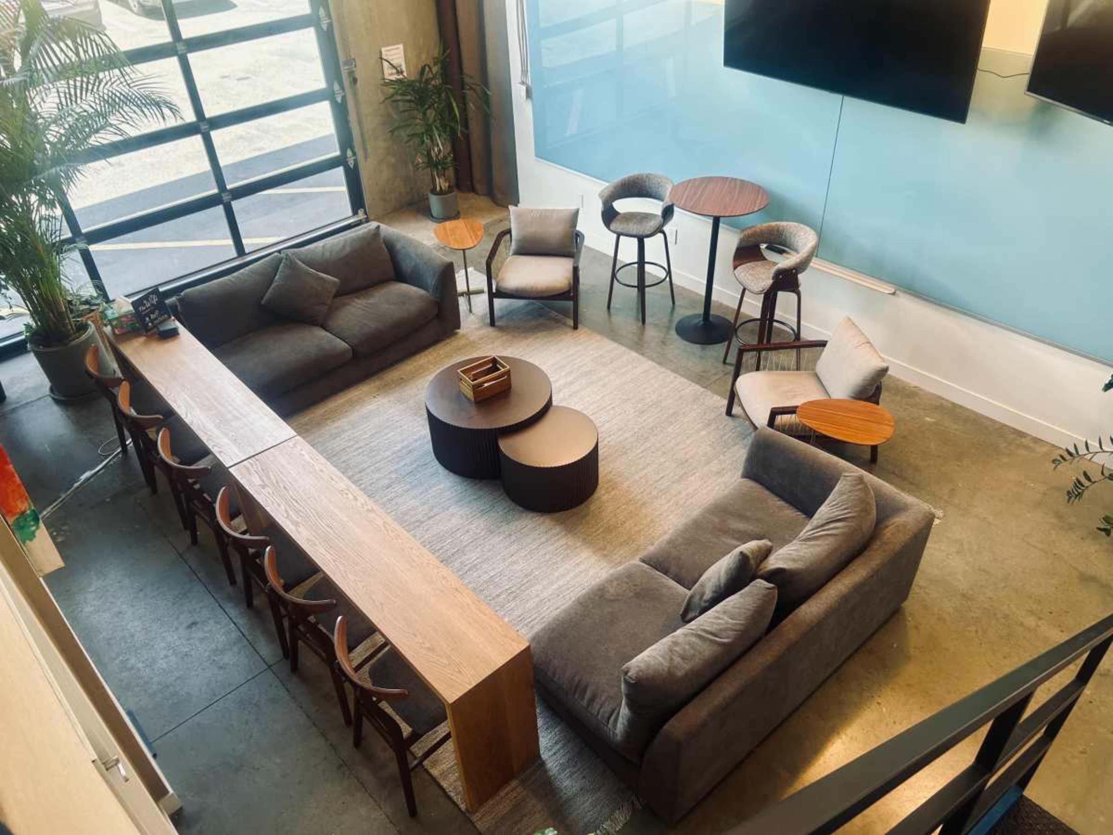 The image shows a modern lounge area featuring two gray sofas, several chairs, a coffee table, and a long wooden table with stools, all arranged on a light-colored rug.