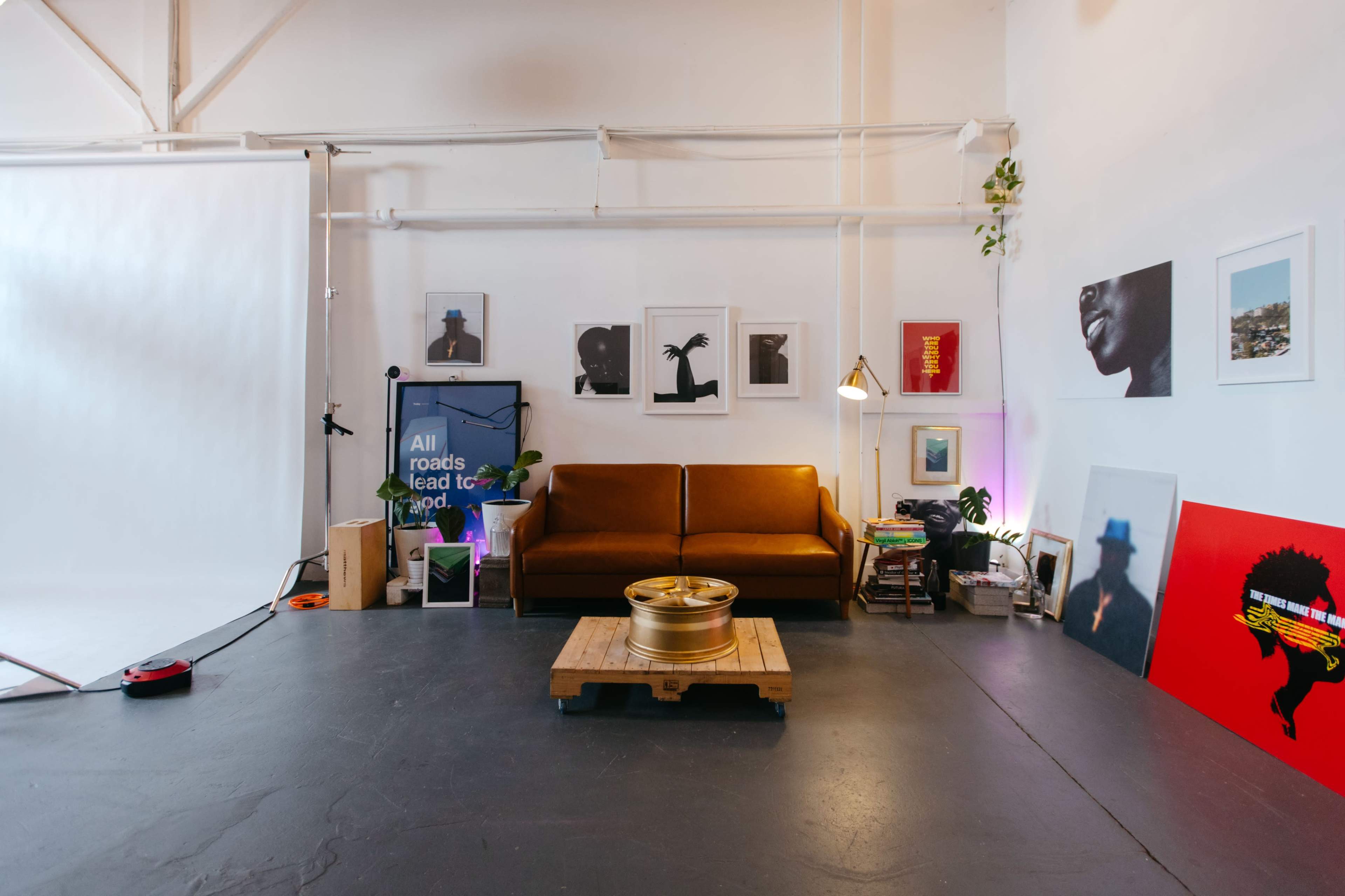 The image shows a loft-style space with a brown sofa, various framed artworks on the walls, and a wooden pallet table in the center.