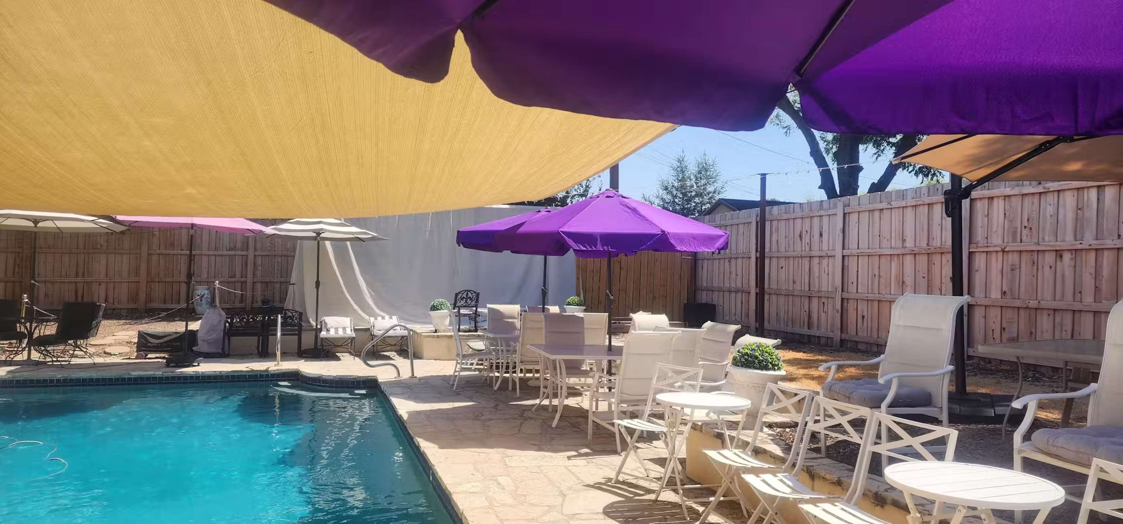 A swimming pool area features purple umbrellas and white patio furniture arranged near the water.