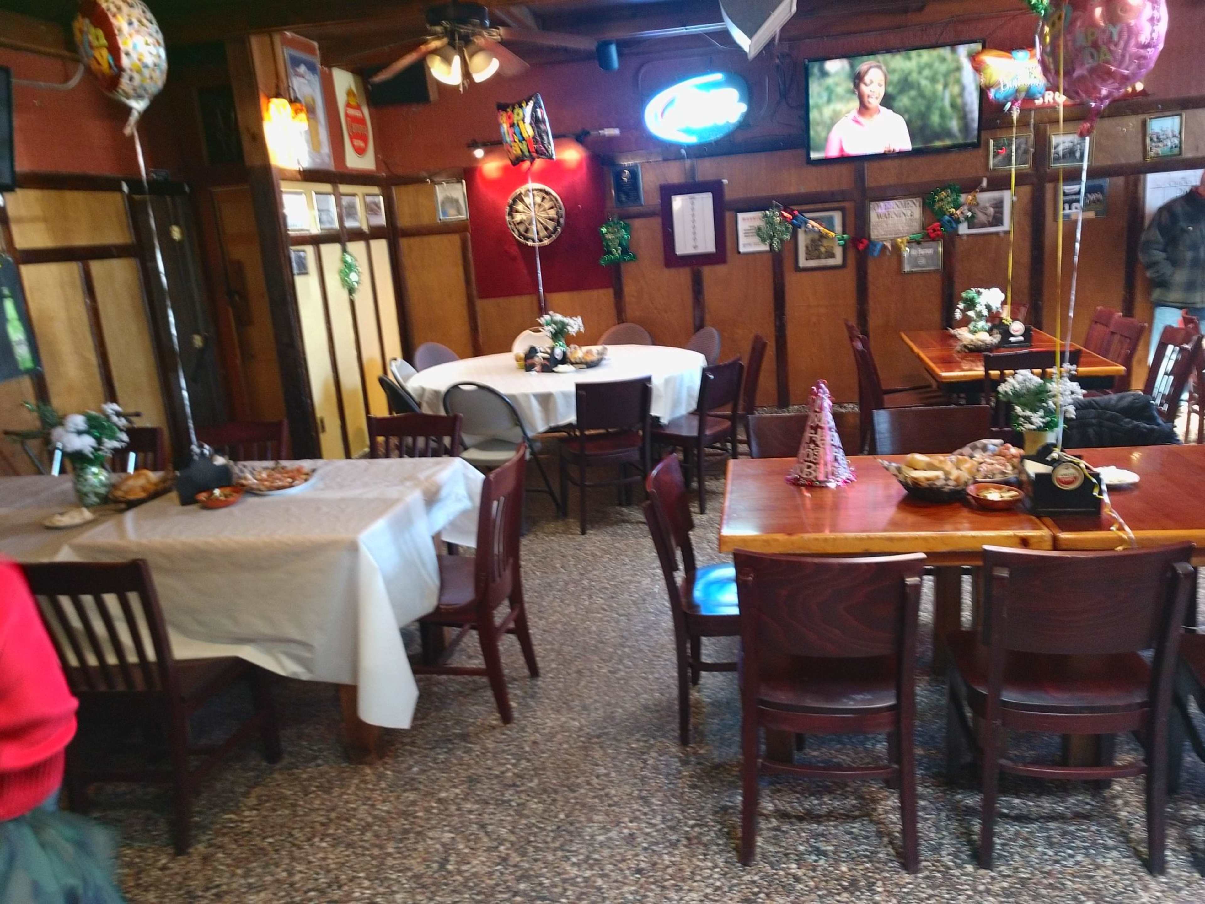 A cozy restaurant interior with wooden tables, chairs, and festive decorations, including balloons and a television screen.