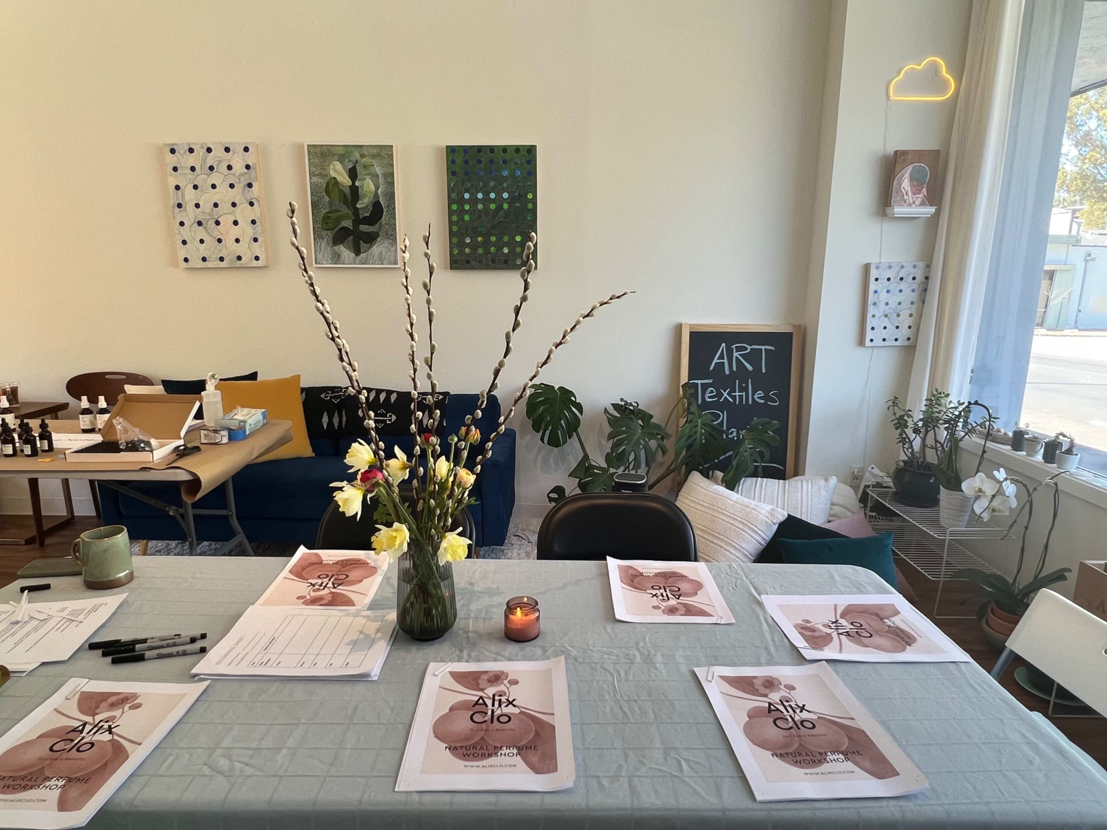 A table with printed materials and a vase of flowers is positioned in a brightly lit room adorned with artwork on the walls and various seating options.