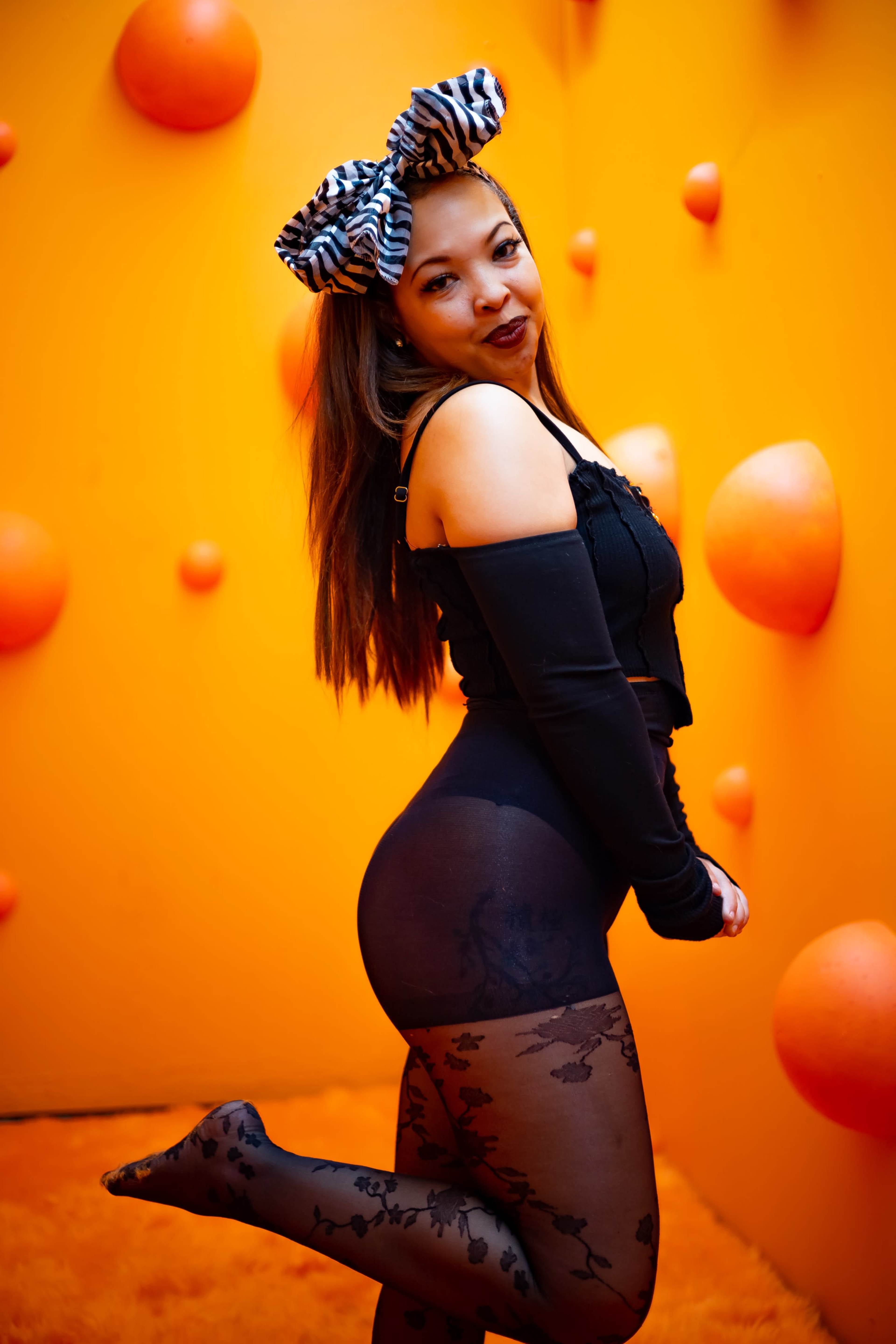 A woman in a black off-shoulder top and patterned tights poses playfully against an orange backdrop with textured decorations.