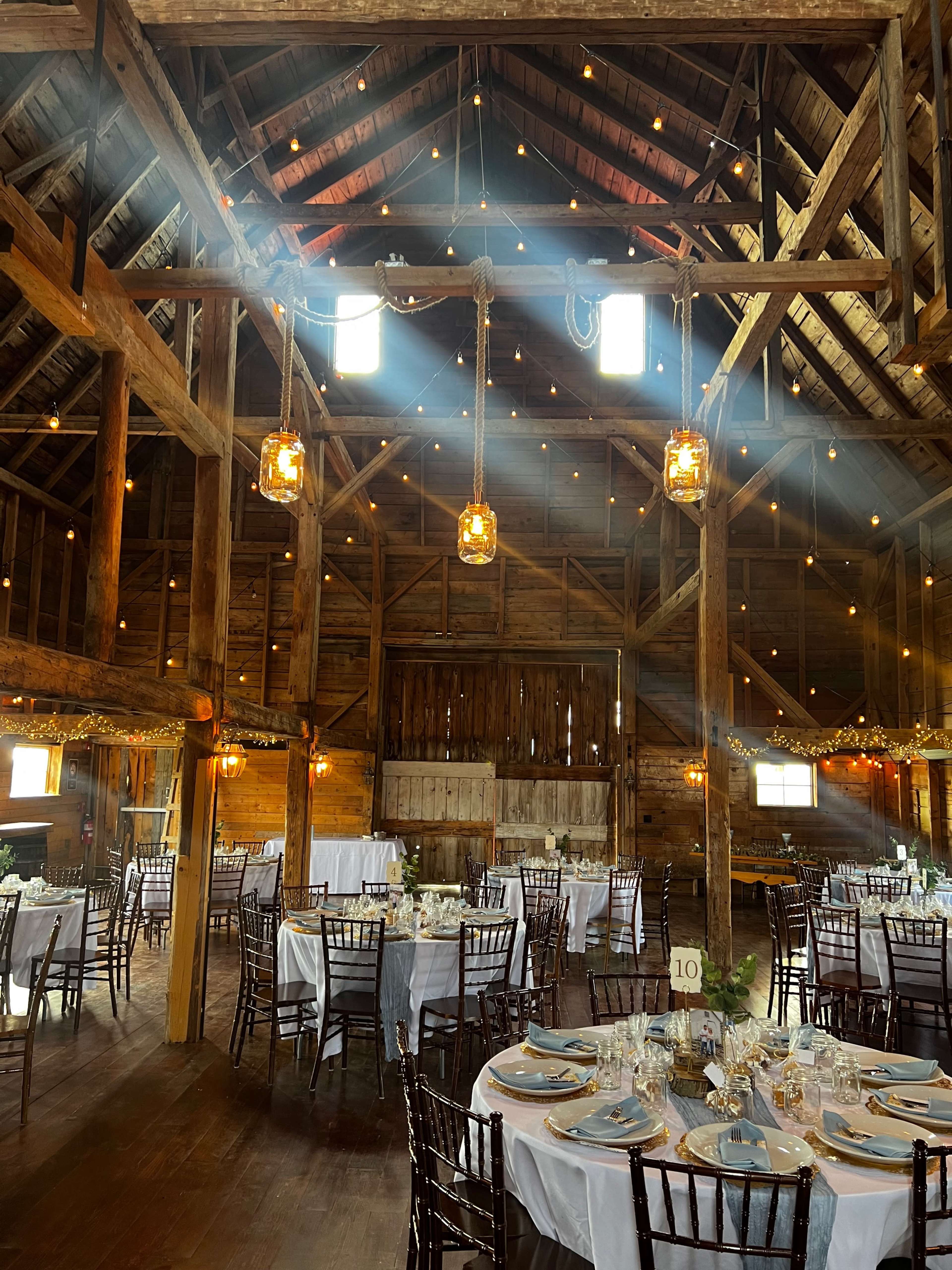 The interior of a rustic barn is set up for a dining event, featuring round tables with white linens, wooden beams, and hanging lanterns.