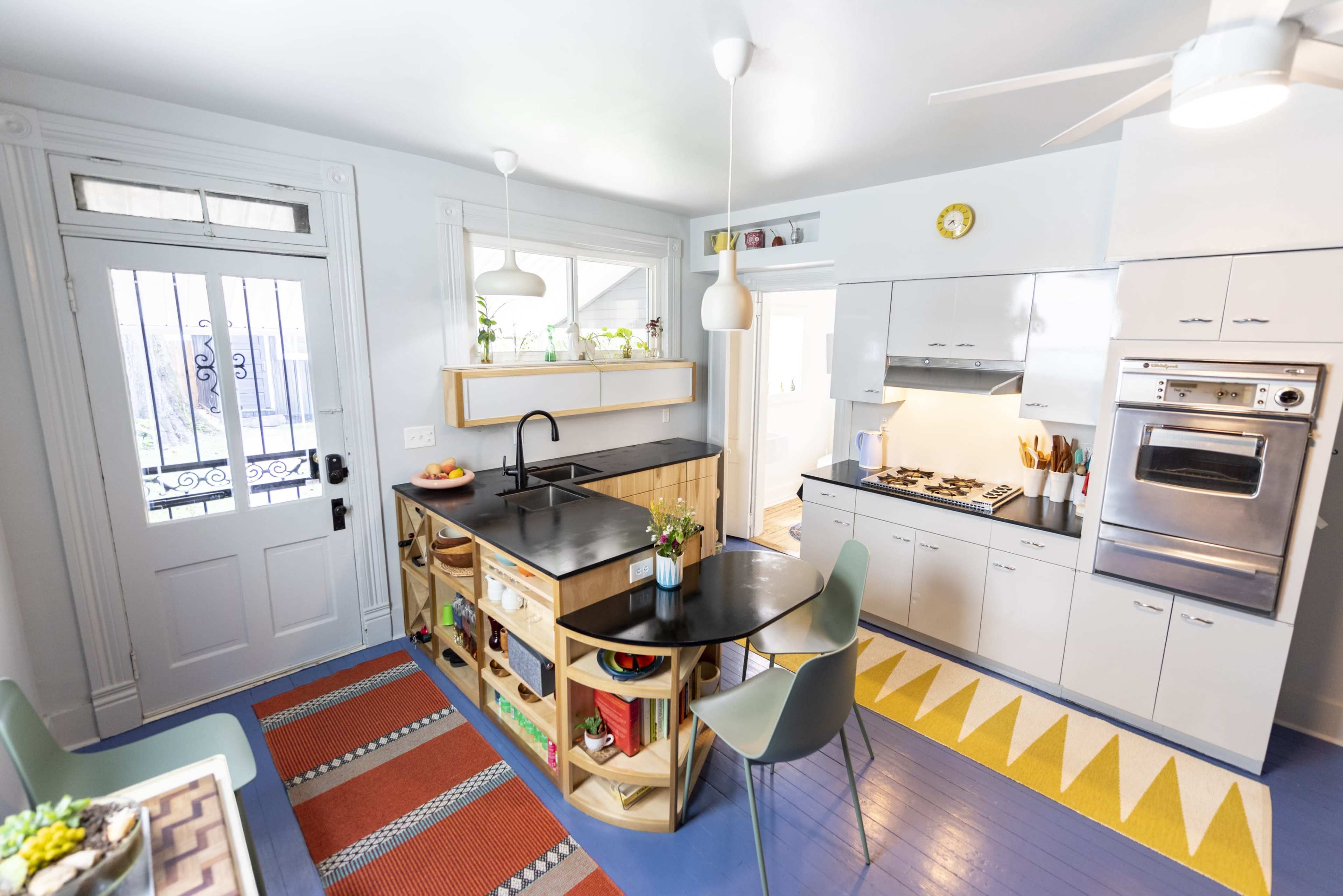 A modern kitchen with a black countertop, open shelving, and colorful flooring, featuring a round table and a door with a decorative grill.