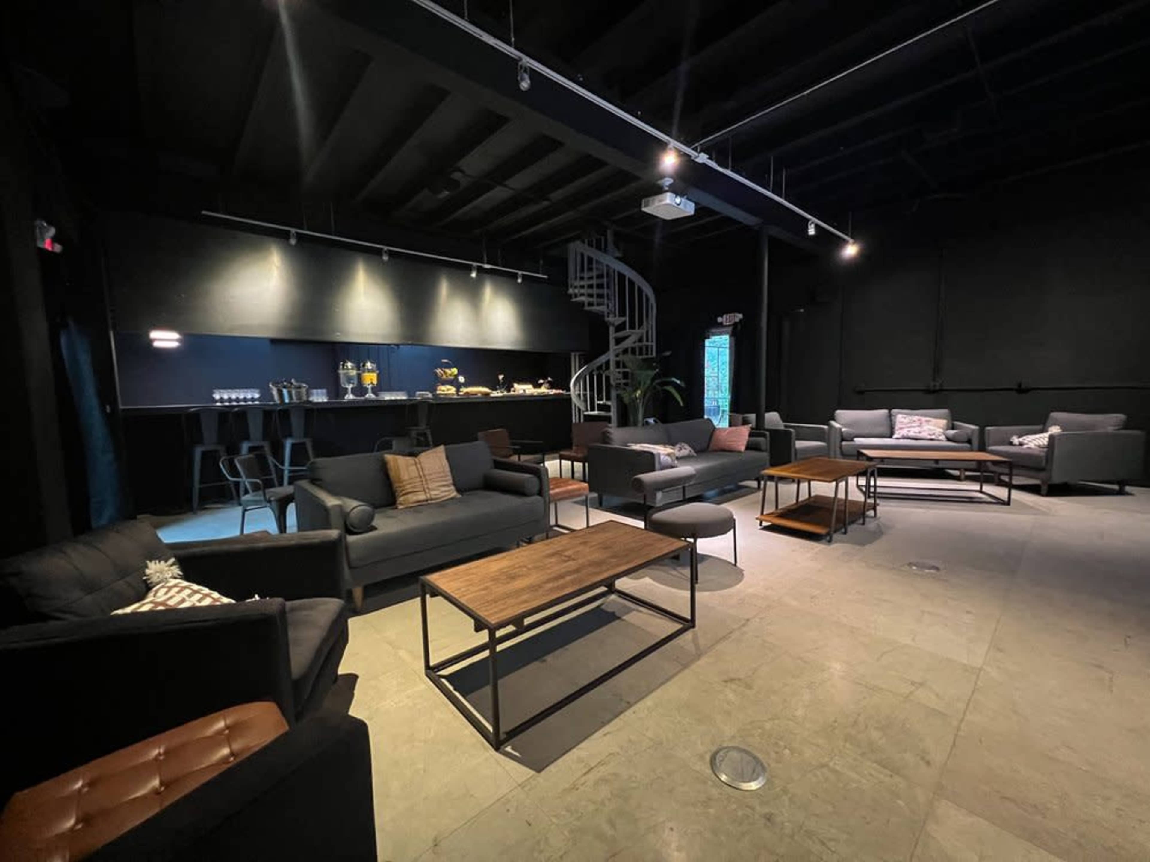 The image shows a modern lounge area with several dark sofas arranged around wooden coffee tables, complemented by a sleek bar in the background.