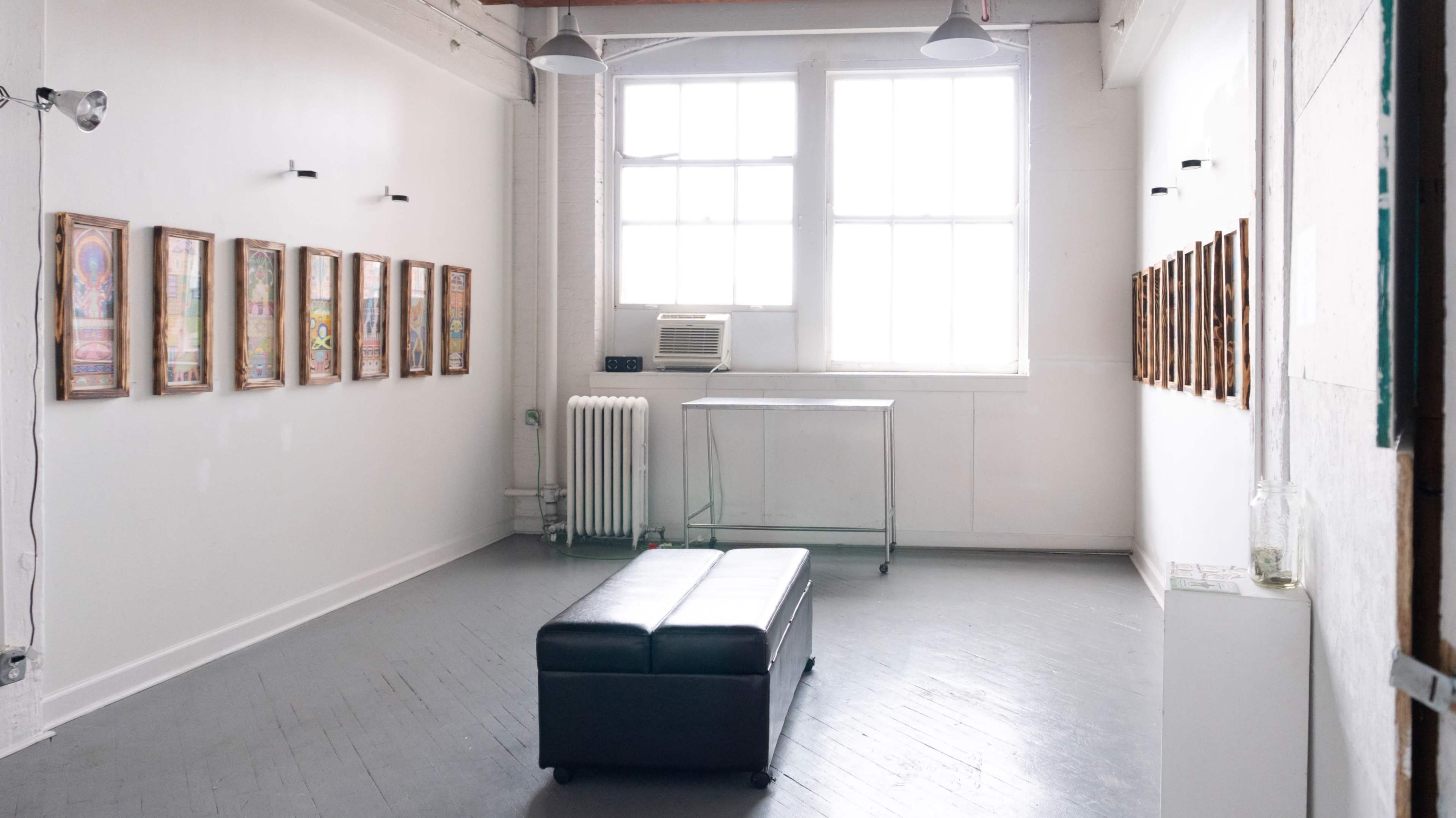 The image shows a bright, minimalist art gallery with framed artworks hanging on the white walls and a black bench in the center.