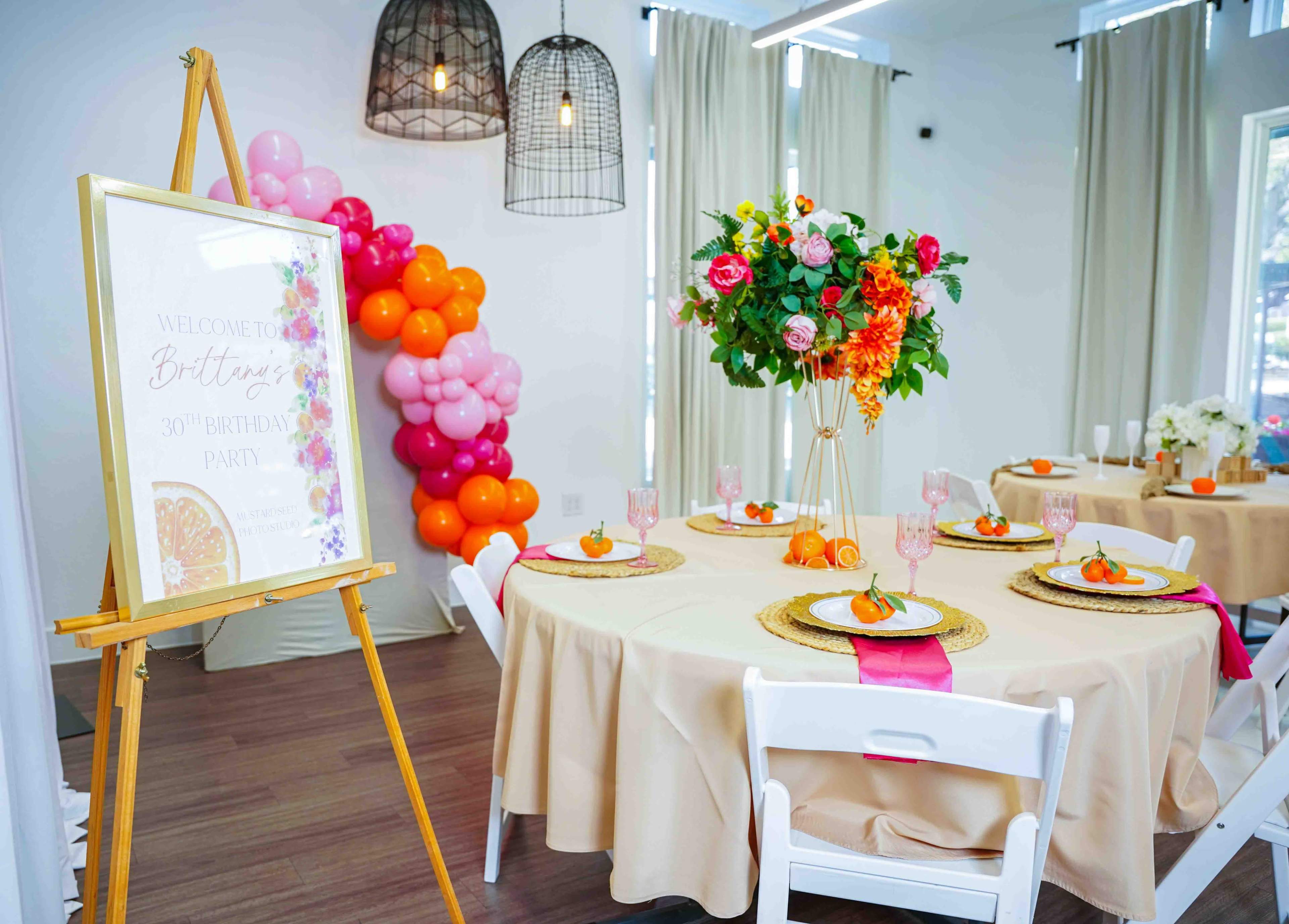 The image shows a decorated party space with a table set for a birthday celebration, featuring a floral centerpiece, colorful balloons, and a welcome sign.