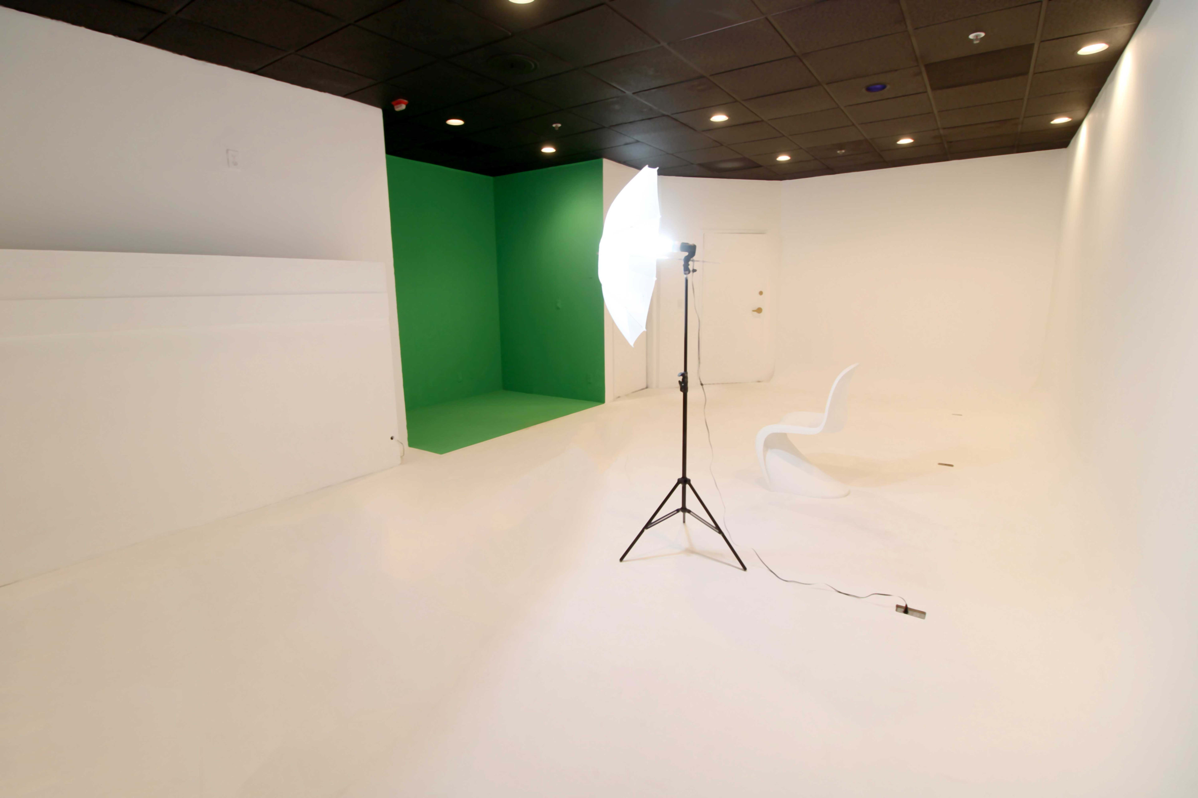 The image shows a photography studio with a white floor, white walls, a green backdrop, and a light stand with an umbrella next to a simple white chair.