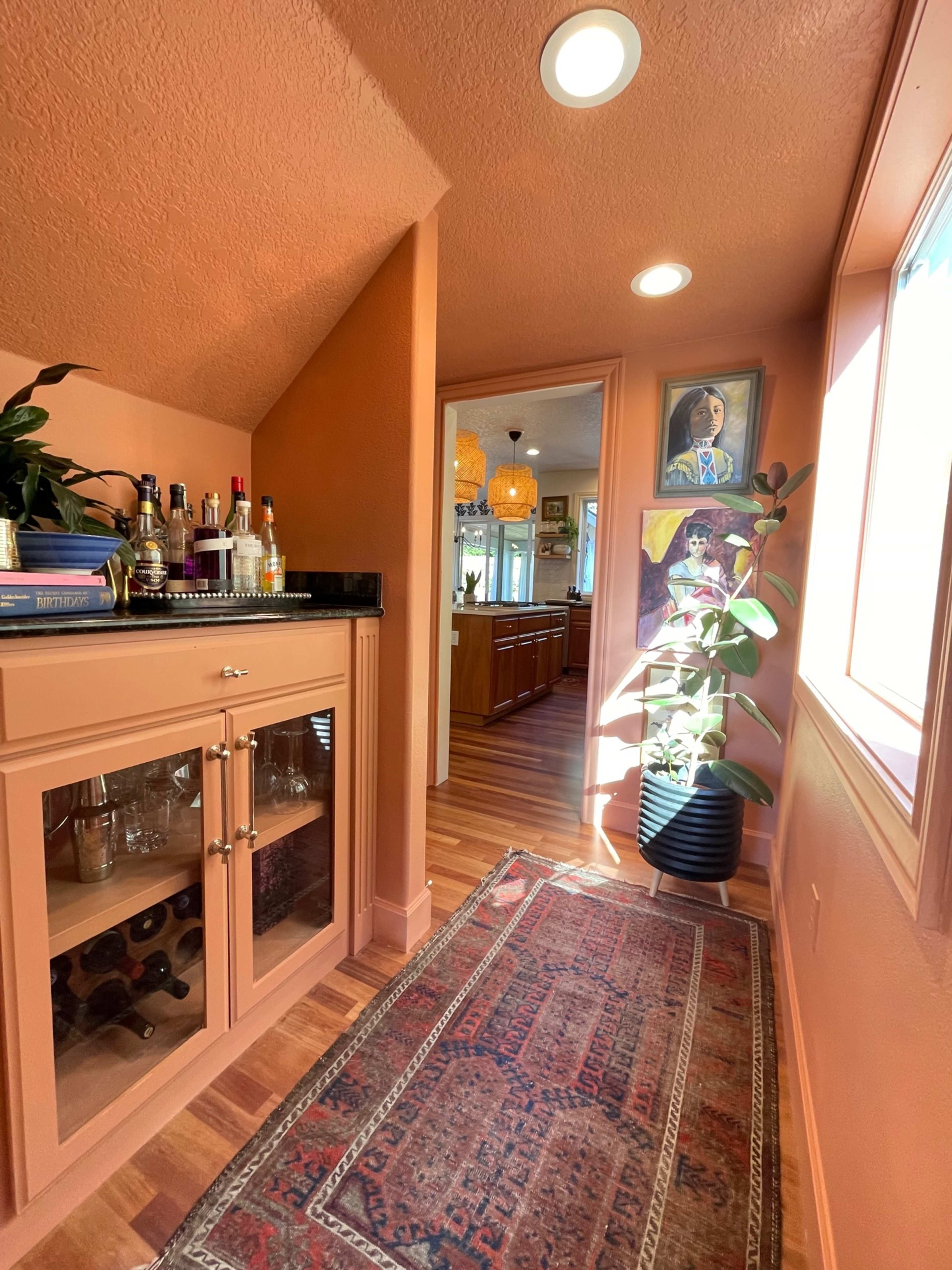 A narrow hallway features a bar cabinet with glassware, a potted plant, and artwork on the walls, leading to a kitchen area beyond.
