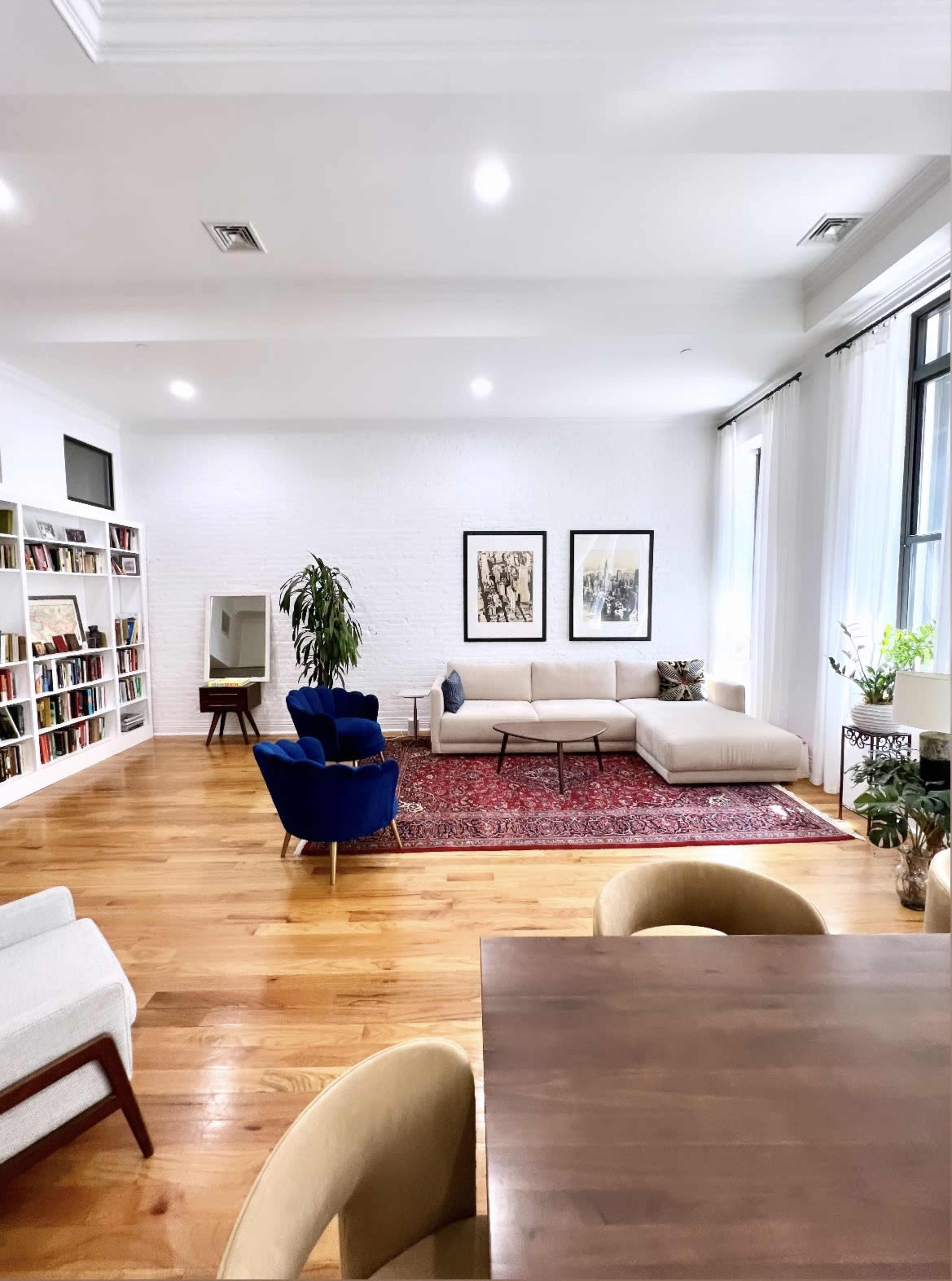A spacious living area featuring a beige sectional sofa, a dark blue accent chair, a wooden dining table, and bookshelves filled with books against a white wall.