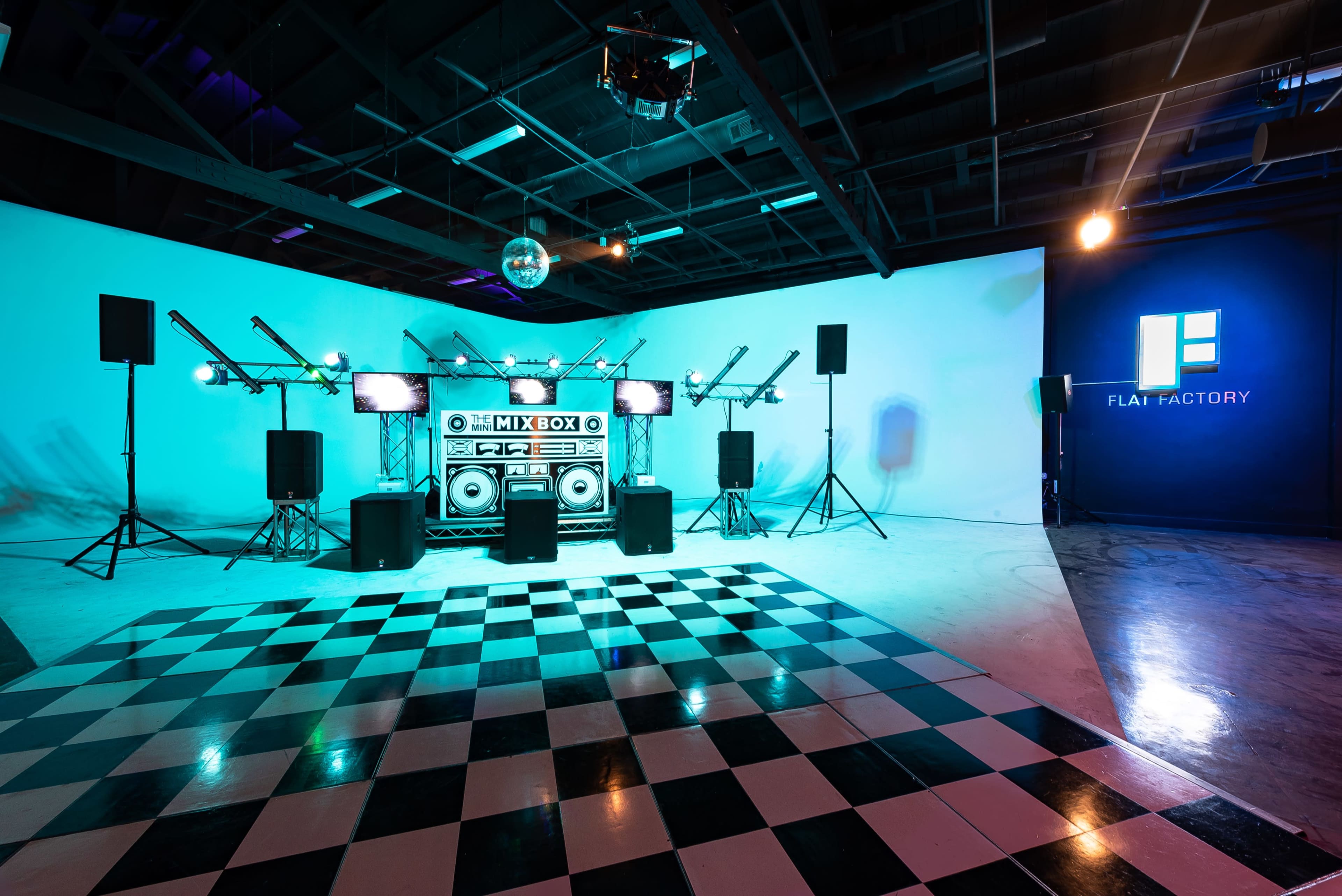 The image shows a music event setup with a checkered dance floor, large speakers, a central DJ booth, and stage lighting in a spacious room featuring a branded wall.