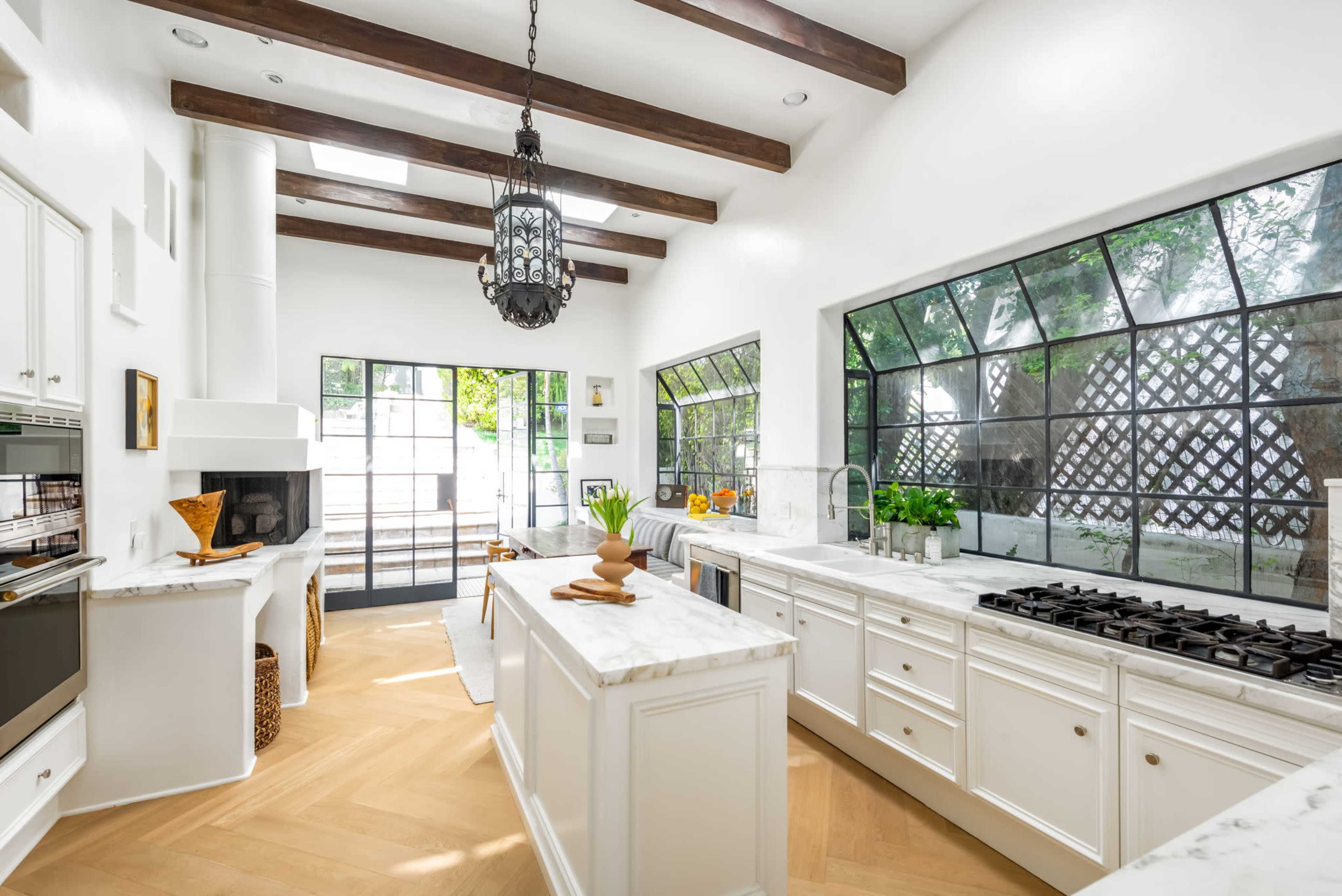 The image shows a modern kitchen featuring white cabinetry, a marble island, large windows with black framing, and exposed wooden beams on the ceiling.