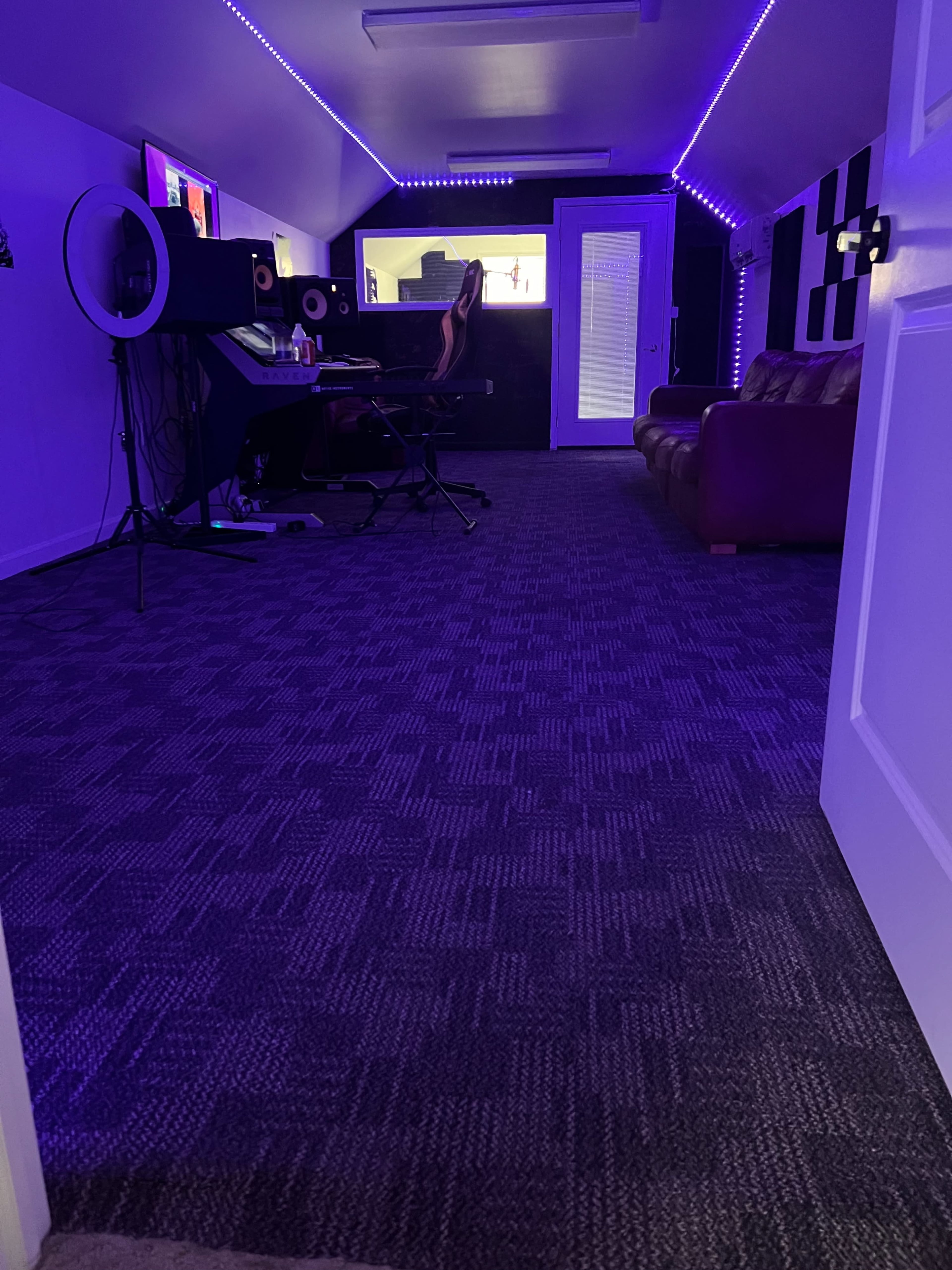 A room with purple lighting features a computer setup, a microphone, and a brown couch, all placed on a carpeted floor.