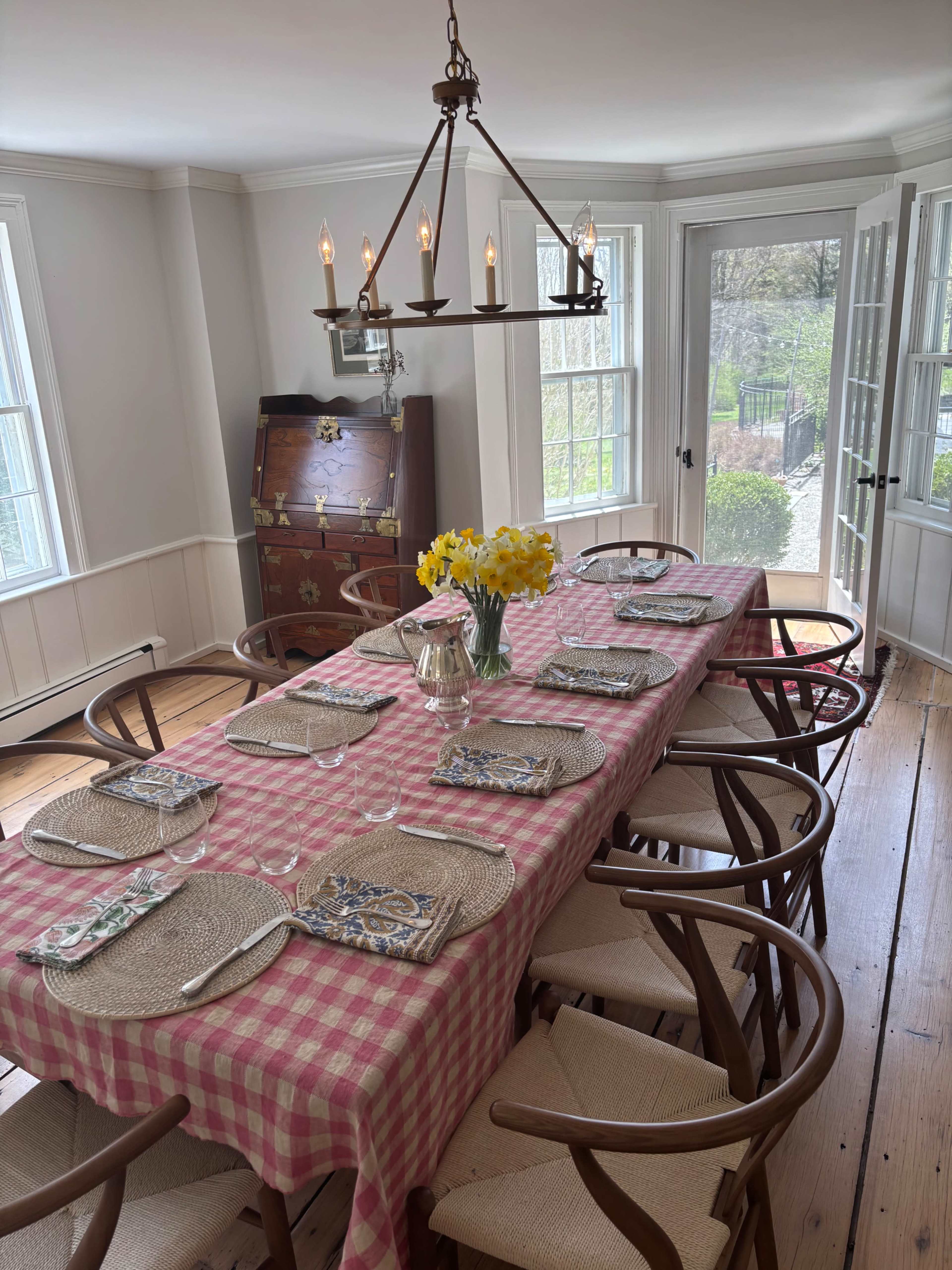 A long dining table is set with plates and utensils, adorned with a checkered pink tablecloth and a vase of yellow flowers, in a well-lit room with large windows.