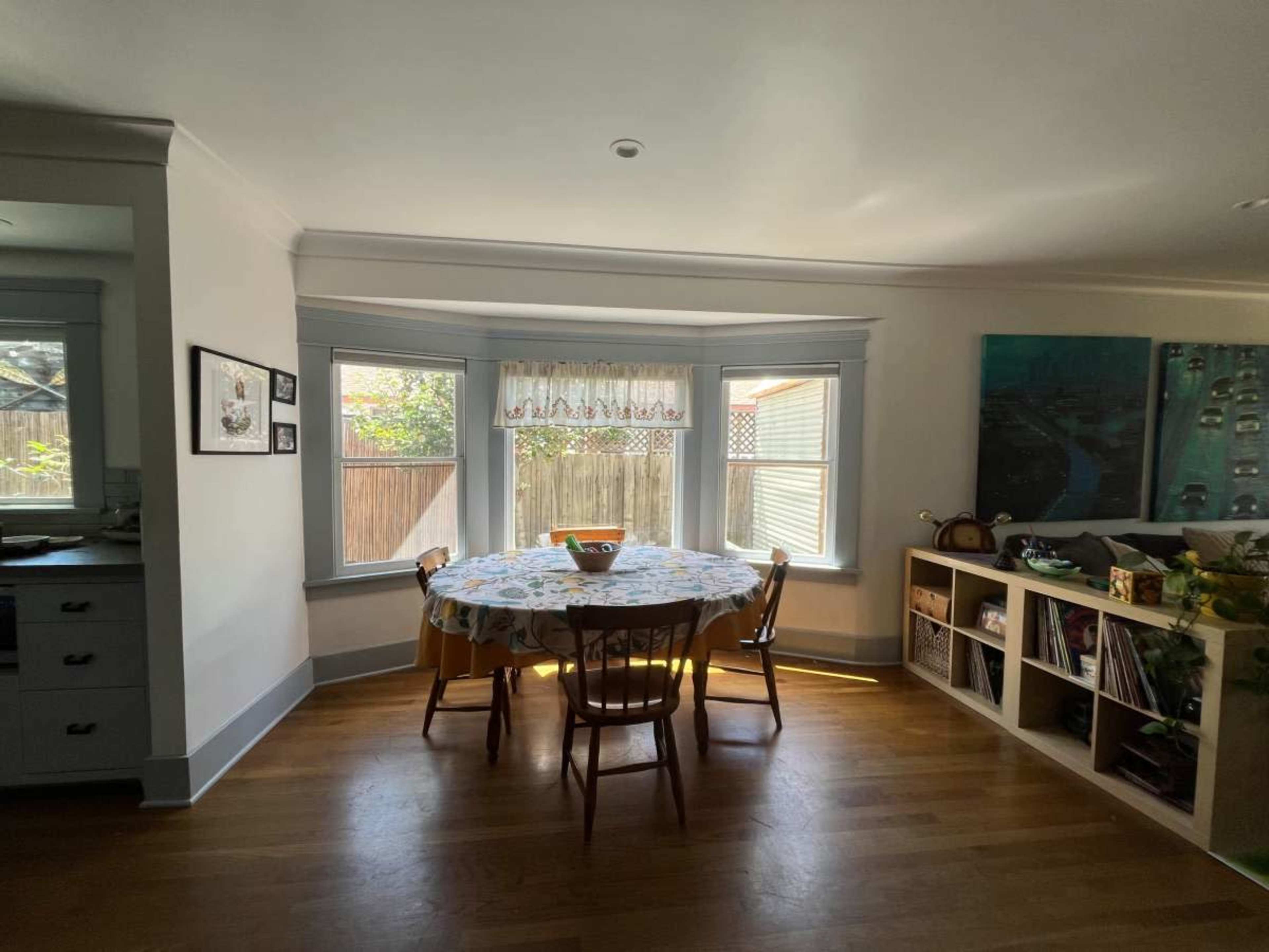 A round table with chairs is positioned near large windows in a bright dining area, with a view of a backyard.