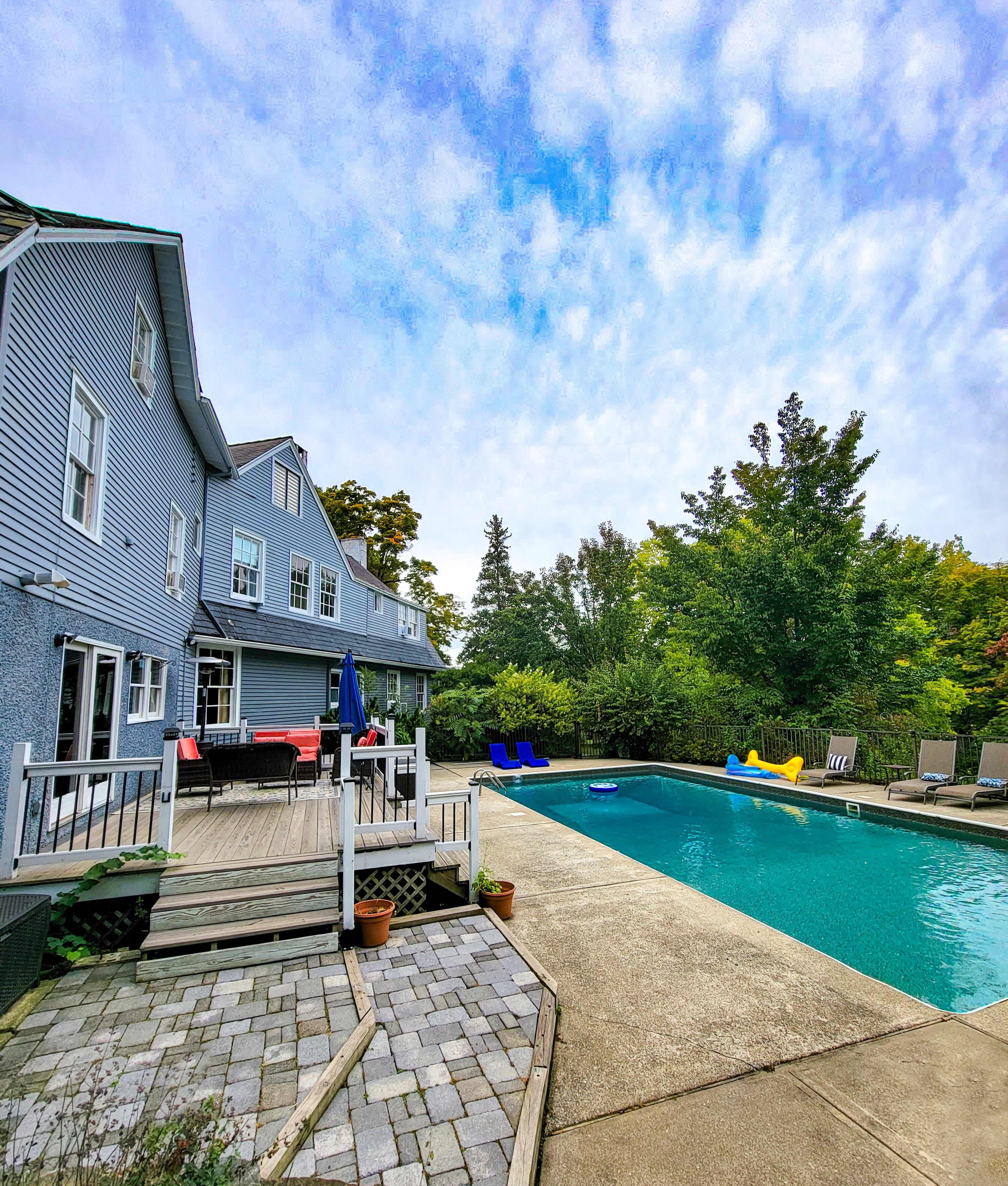 A large rectangular swimming pool is surrounded by a patio, which features lounge chairs and a deck leading to a house with multiple windows.