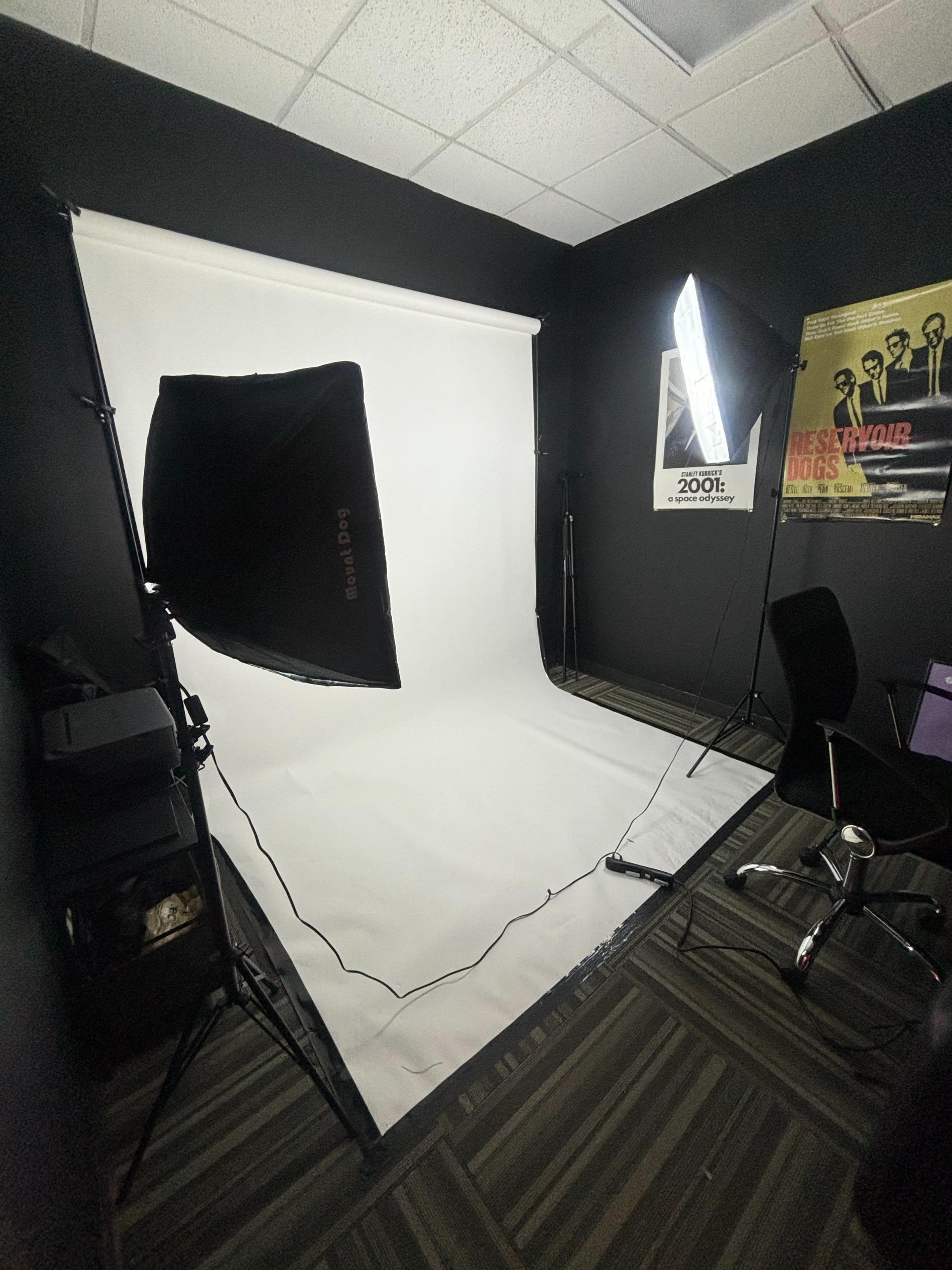 The image shows a photography studio with a white backdrop, studio lights, and a black office chair in a dimly lit room.