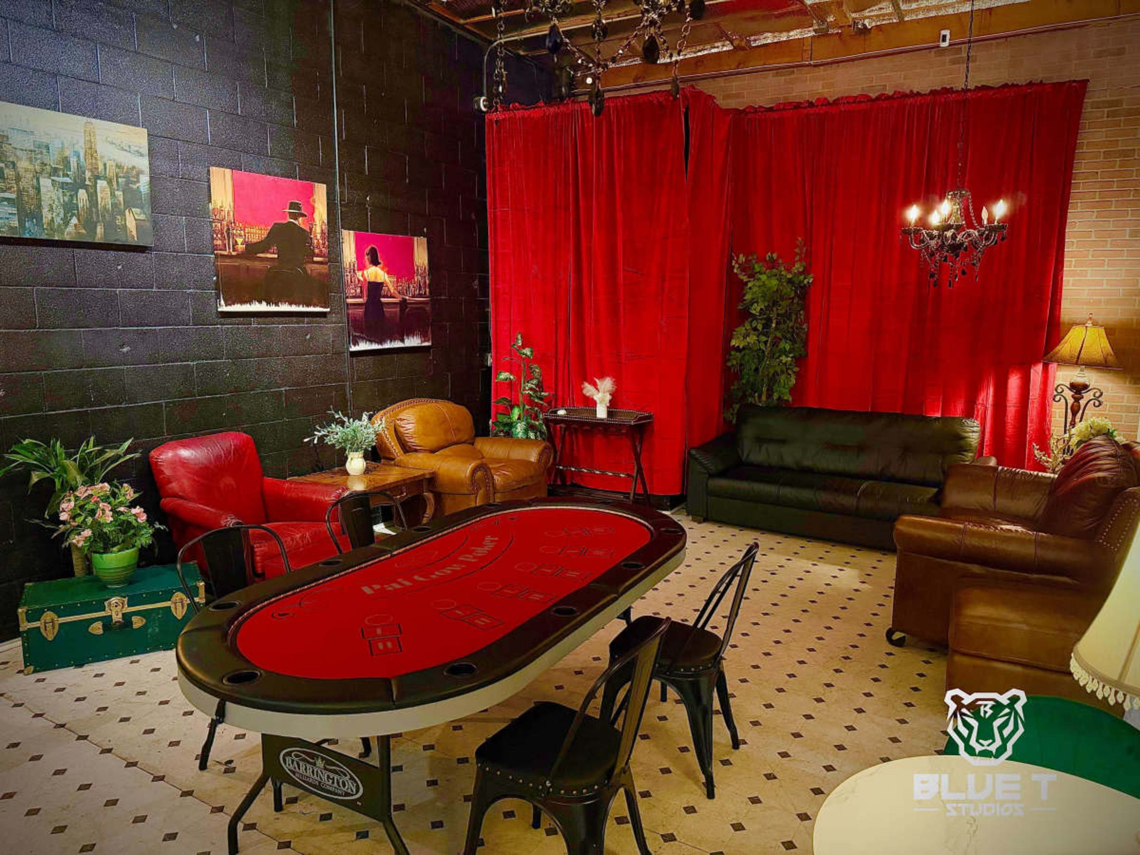 The image features a professionally designed lounge area with a poker table, comfortable seating, a chandelier, and decorative red curtains against a black wall.
