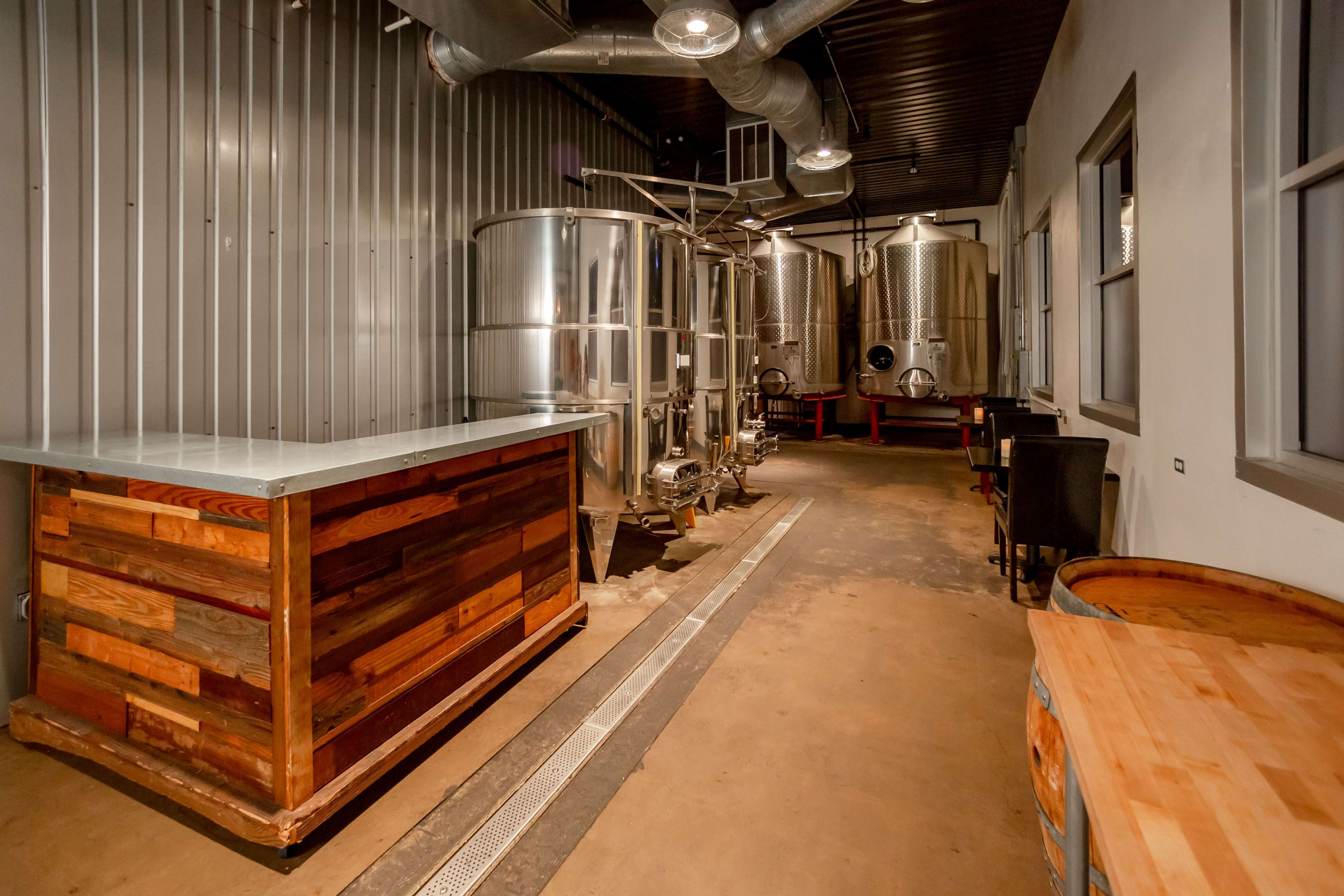The image shows a modern brewing facility with stainless steel fermentation tanks, a wooden serving counter, and an unfinished concrete floor.