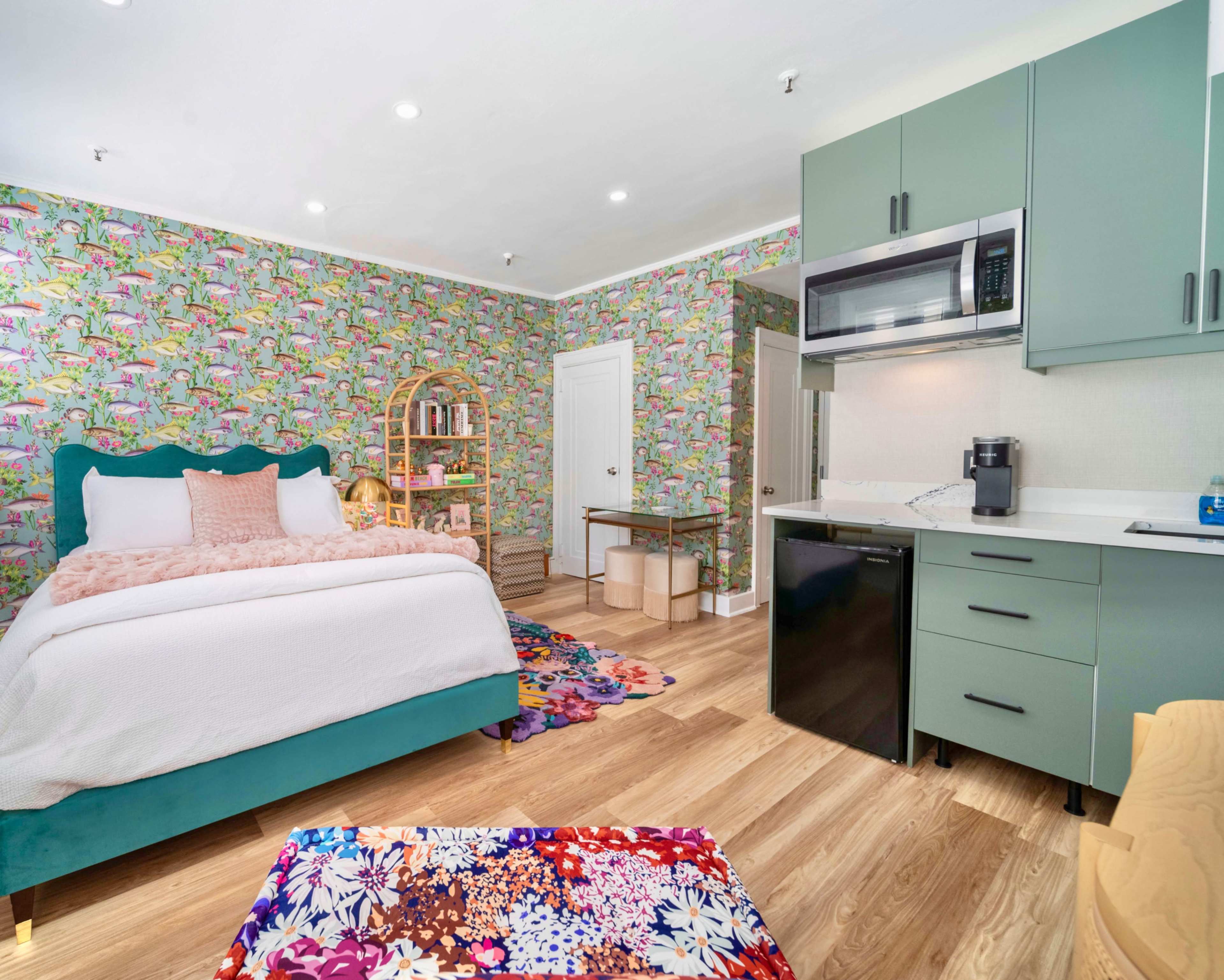 A brightly decorated bedroom featuring a bed with a green headboard, colorful floral wallpaper, and a small kitchen area with a microwave and refrigerator.