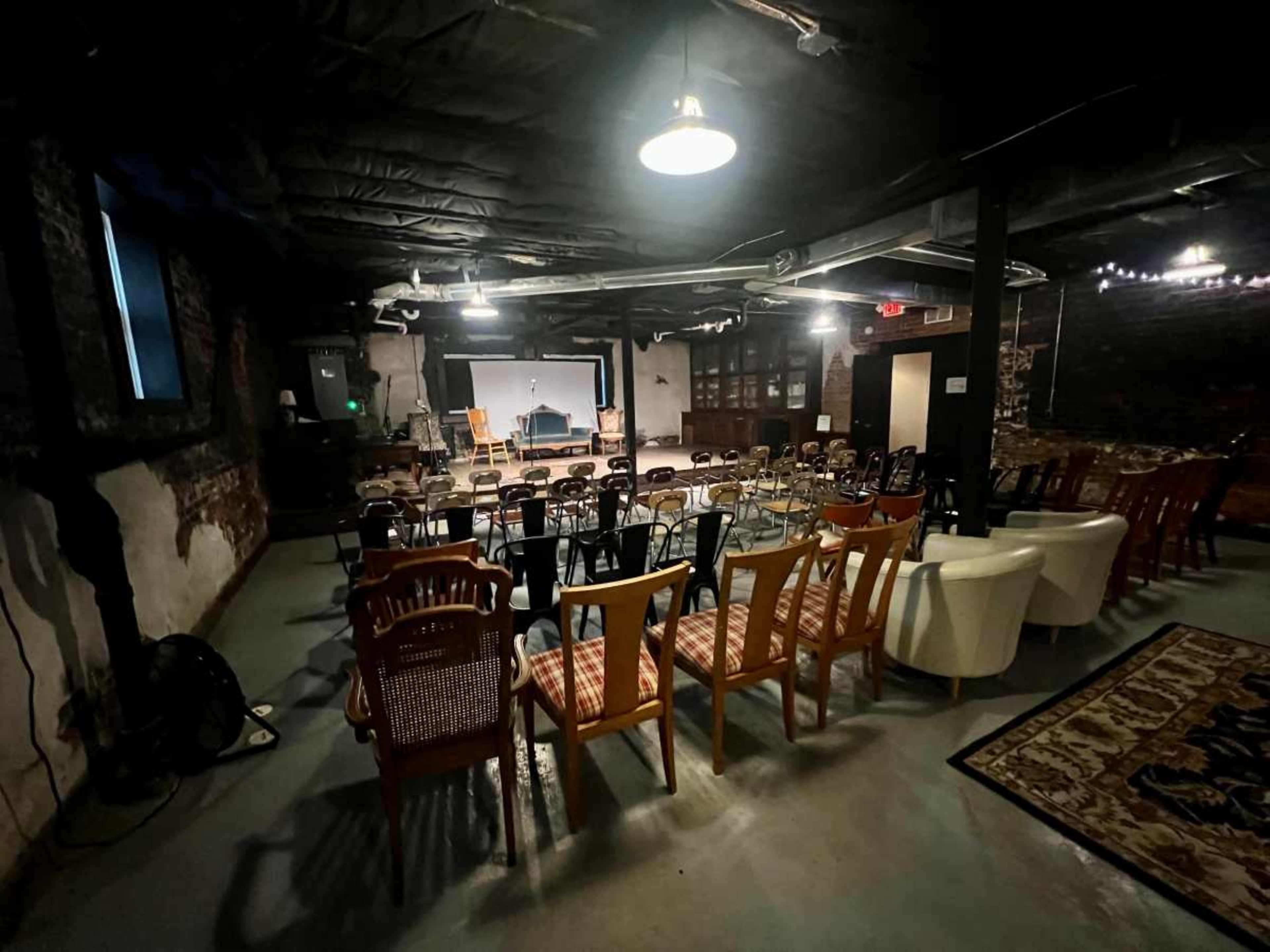 The image shows a dimly lit room with a stage at one end, surrounded by rows of various chairs and a carpet on the floor.