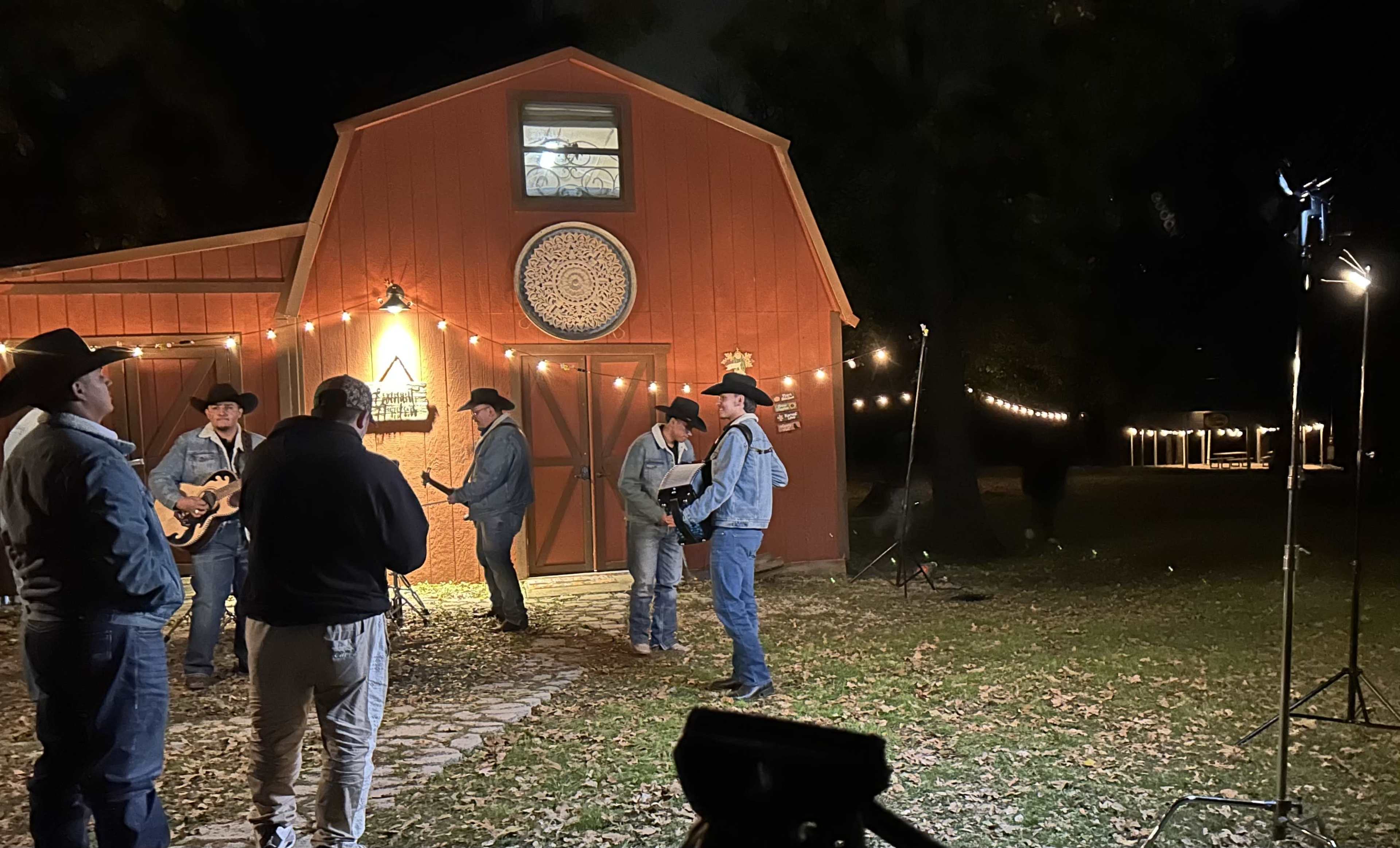 A group of musicians dressed in cowboy attire is performing outside a rustic orange barn illuminated by string lights at night.