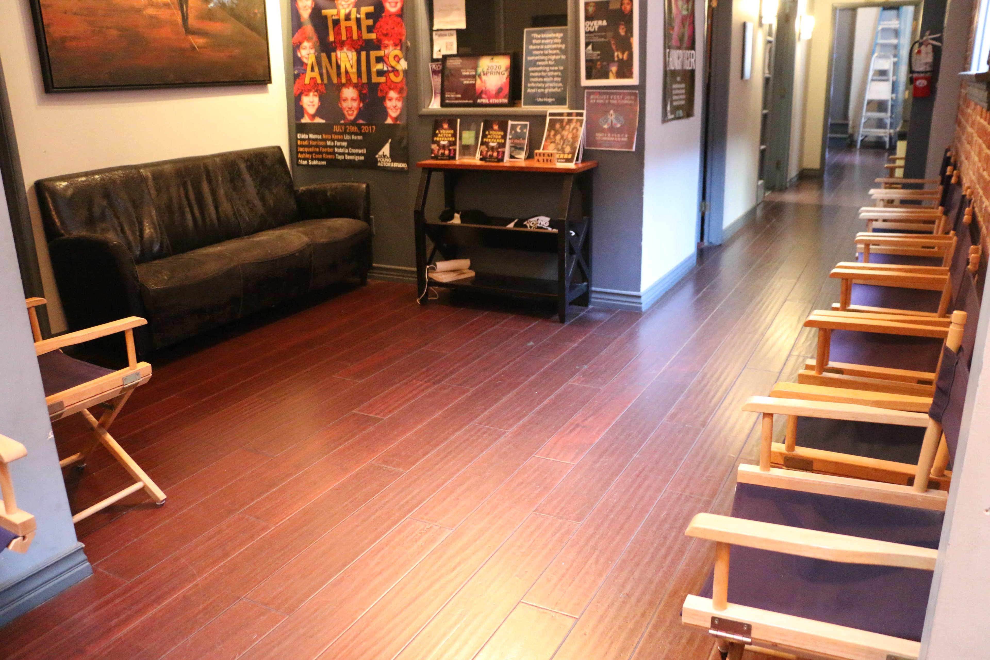 The image shows a corridor with wooden flooring, featuring a black sofa on one side and several wooden chairs arranged along the opposite wall, with promotional posters displayed.