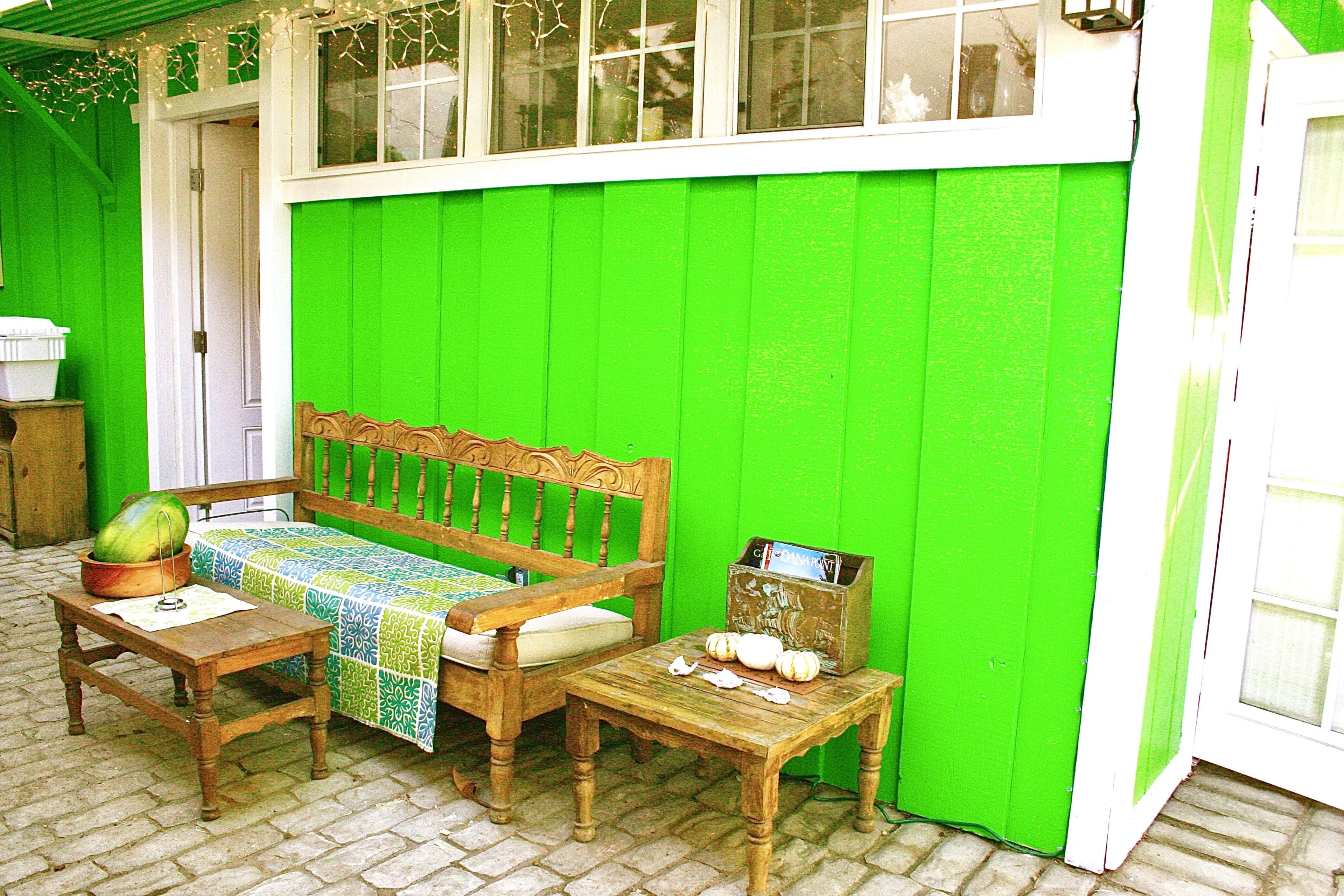 A wooden bench and two tables sit in front of a vibrant green wall, with a patterned cloth draped over the bench and small decorative items on the tables.