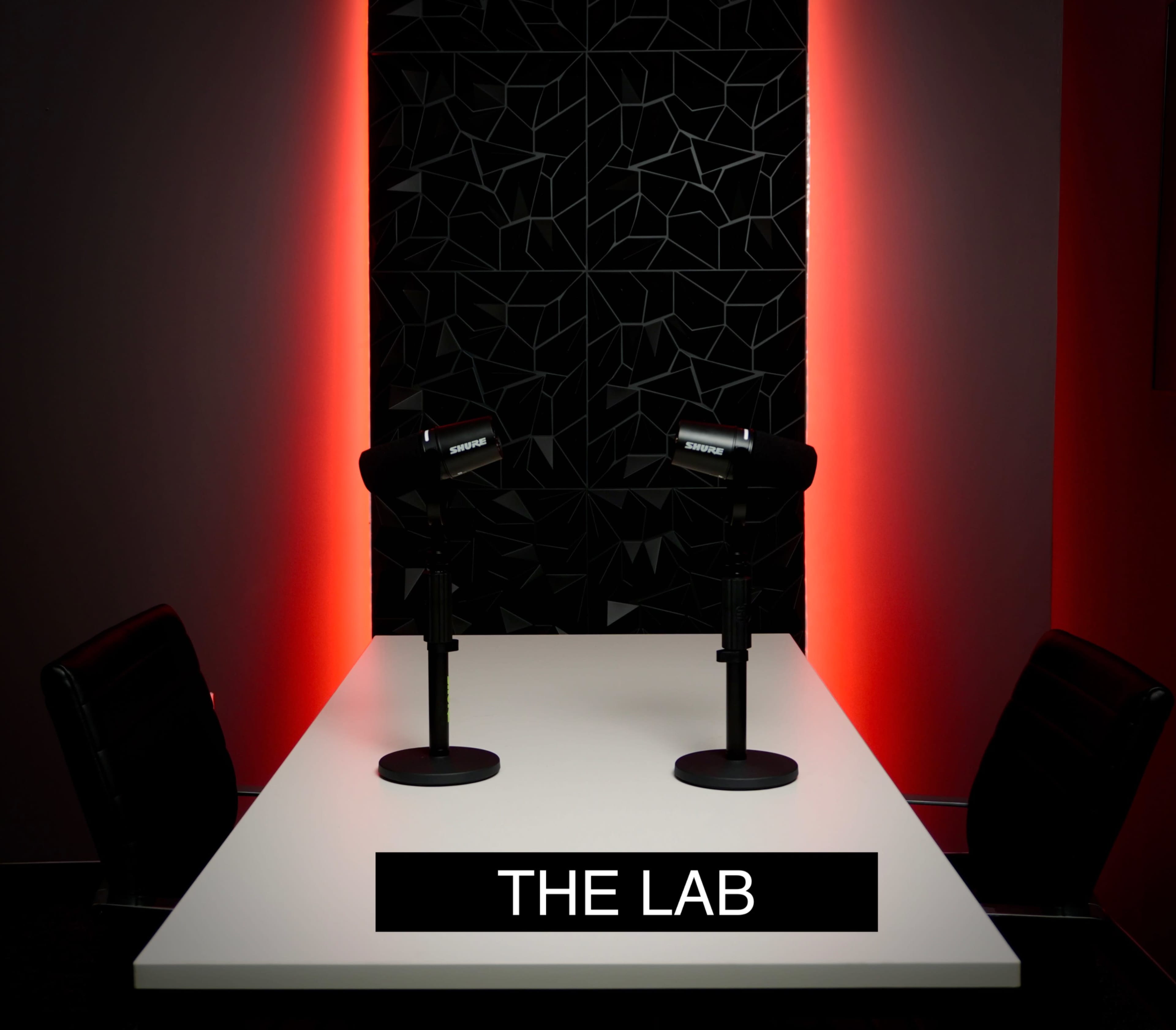 A brightly lit table with two microphones is set against a backdrop featuring geometric patterns and illuminated with red lighting, labeled "THE LAB."