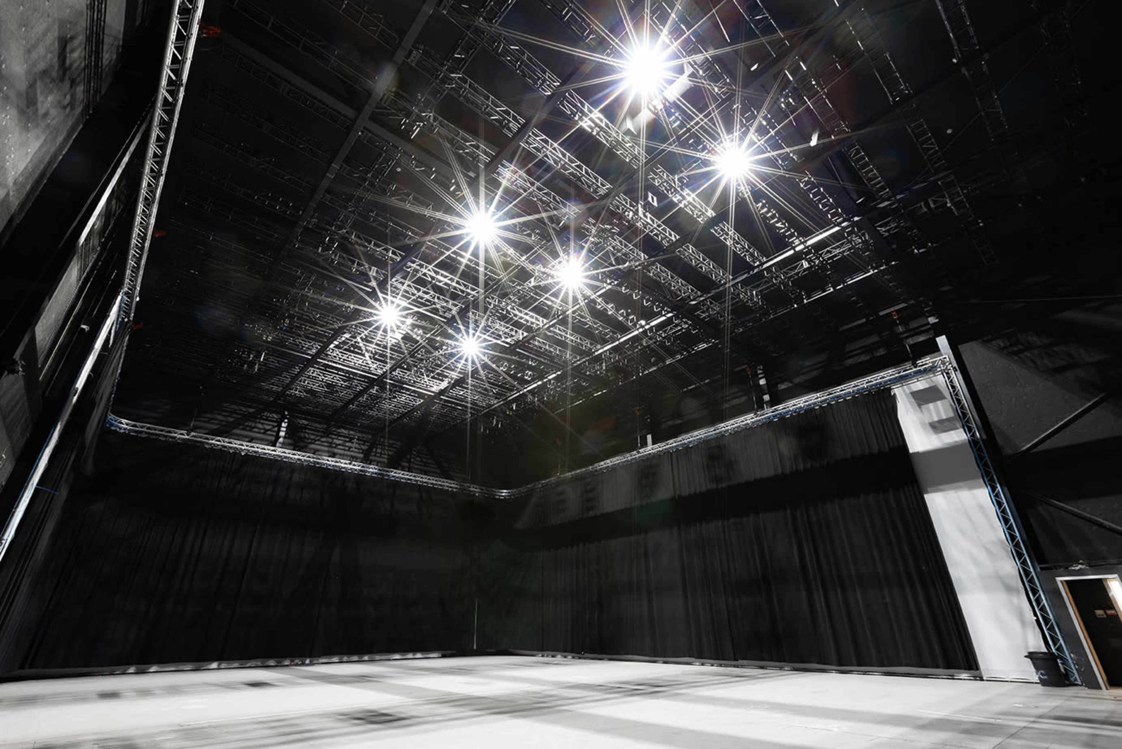 A large, empty stage area is illuminated by bright overhead lights and surrounded by black curtains.